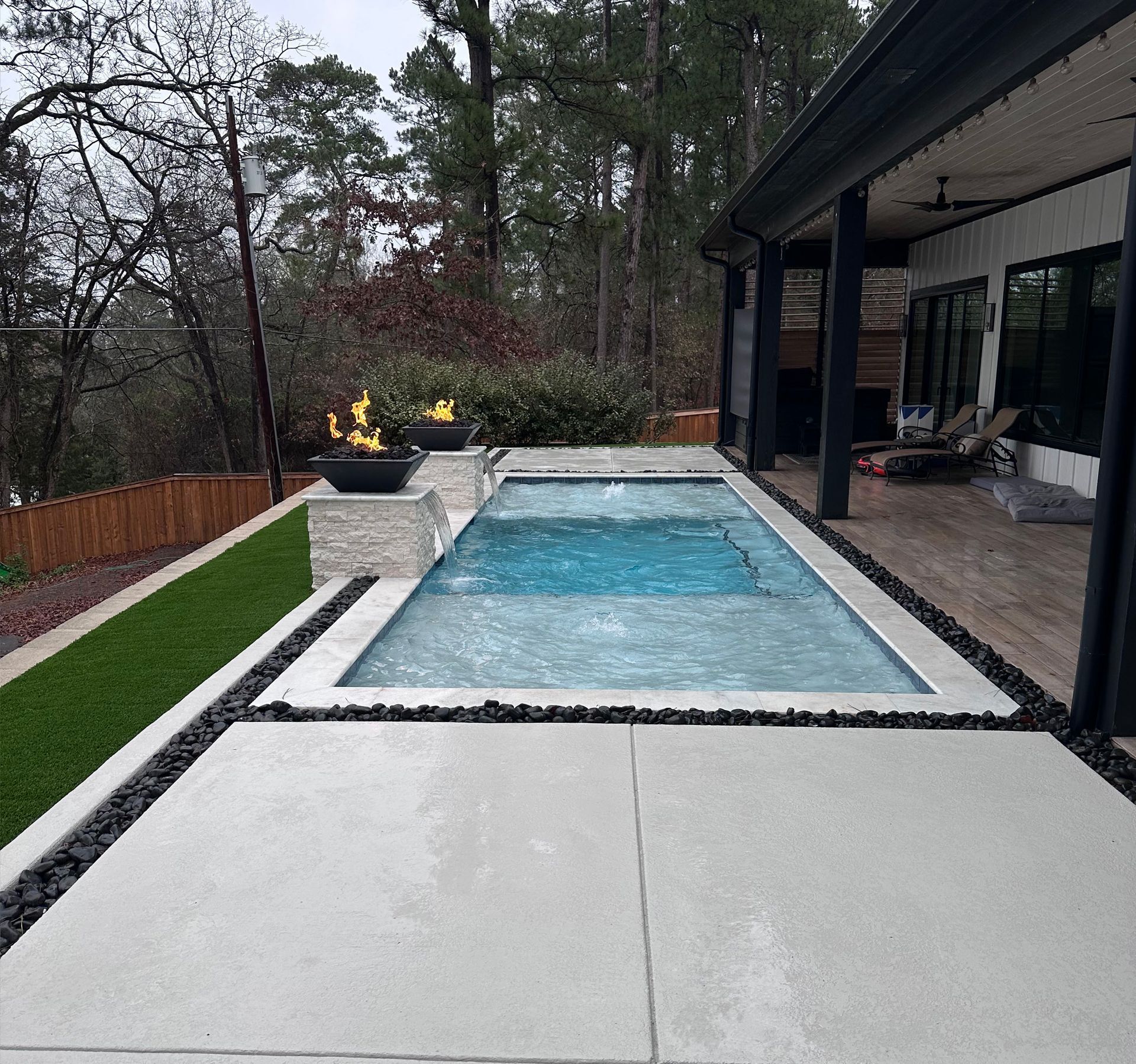 Backyard with a rectangular pool, fire pit, patio, and lush trees.