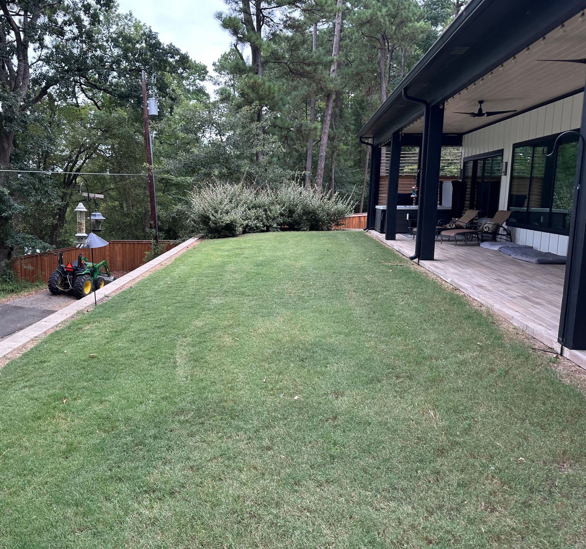 Green lawn beside a porch with a dark roof and black pillars, with trees and a mower in the background.