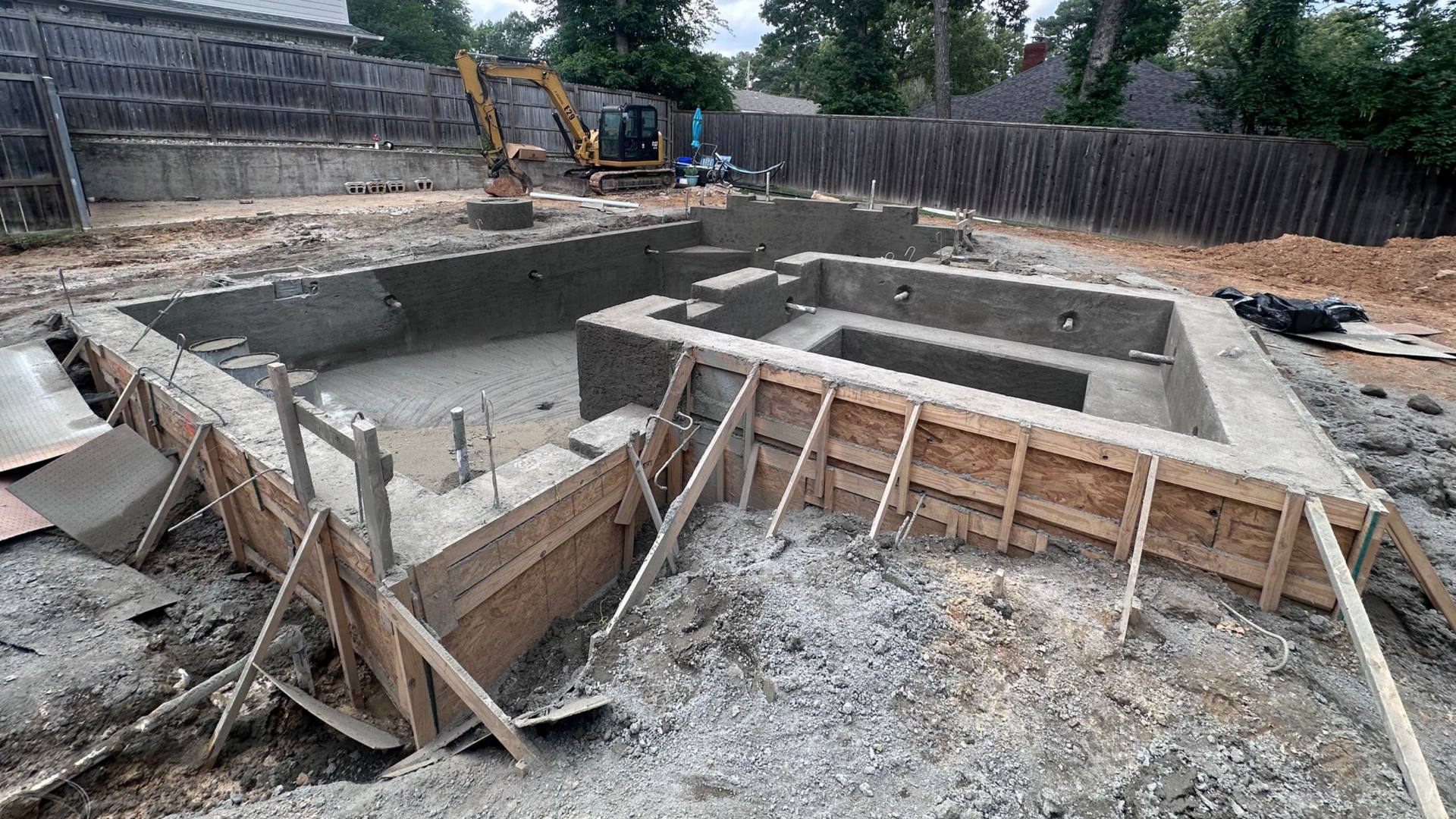 Pool construction site with concrete forms, excavator, and excavated earth.