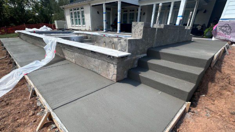 Construction site with concrete patio, steps, and pool foundation next to a white house.