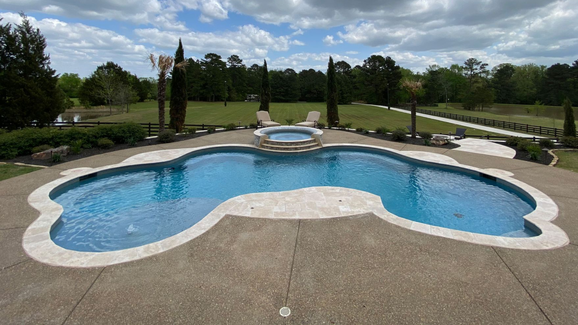 Swimming pool with a connected hot tub, surrounded by a stone patio. Green lawn and trees in the background.