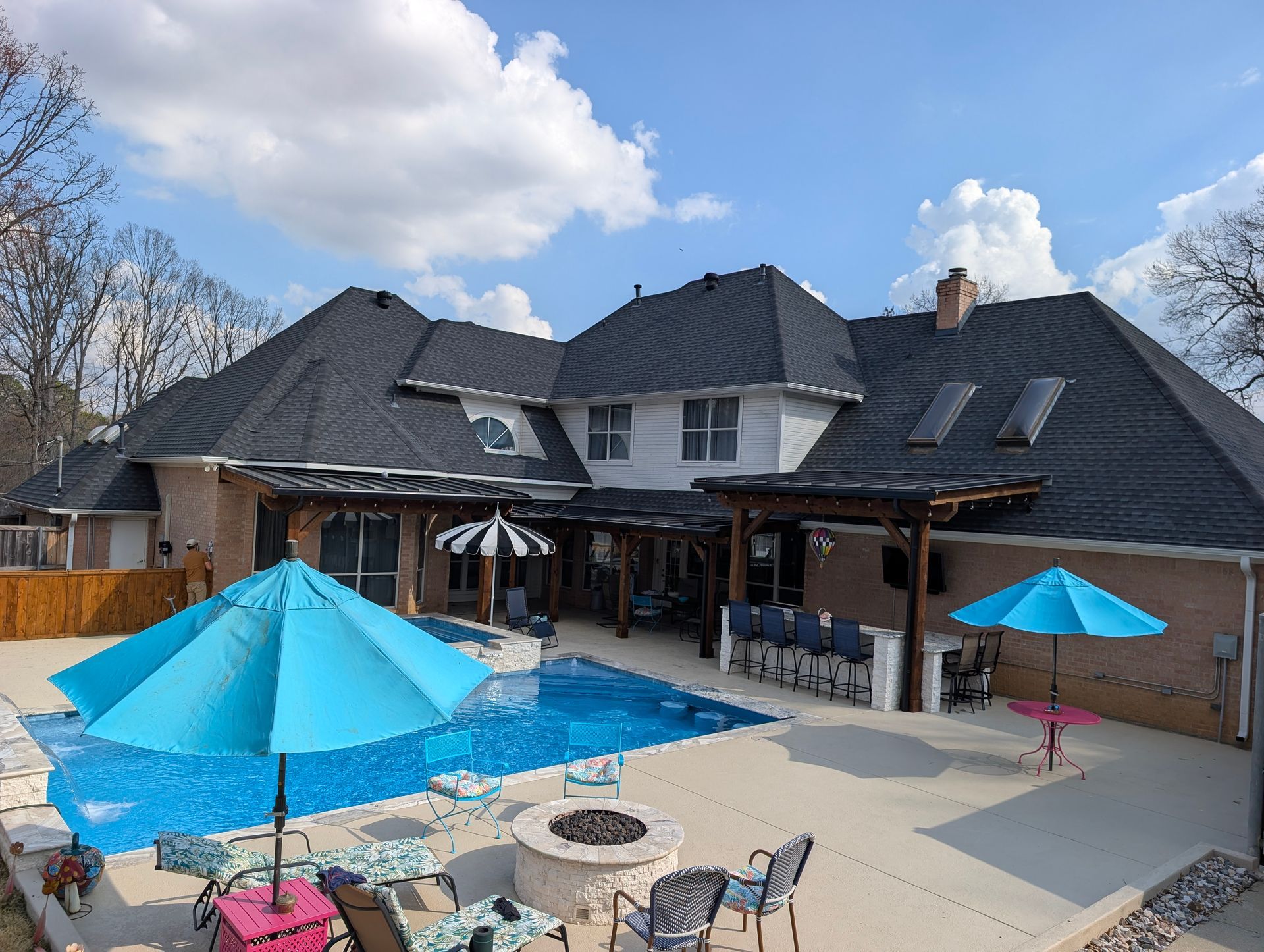 Backyard with a pool, patio, and house. Blue umbrellas, lounge chairs, and a fire pit are visible. Sunny day.