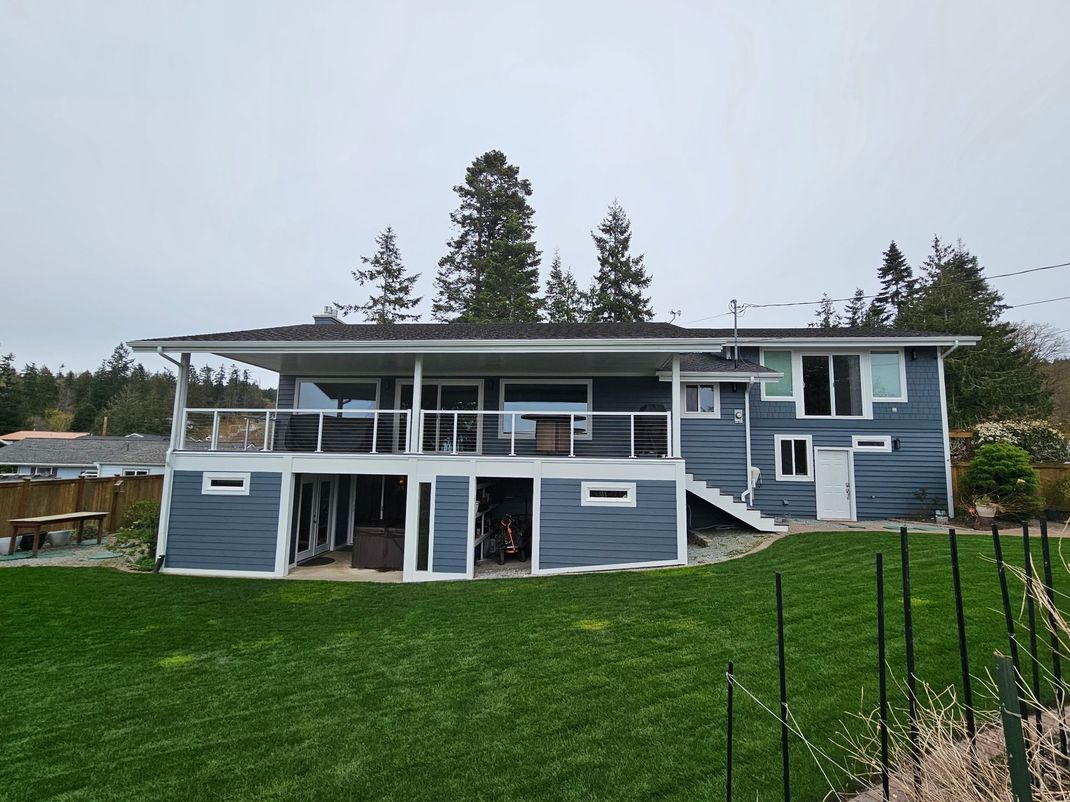 Blue two-story house with white trim and large deck on a grassy hillside under an overcast sky