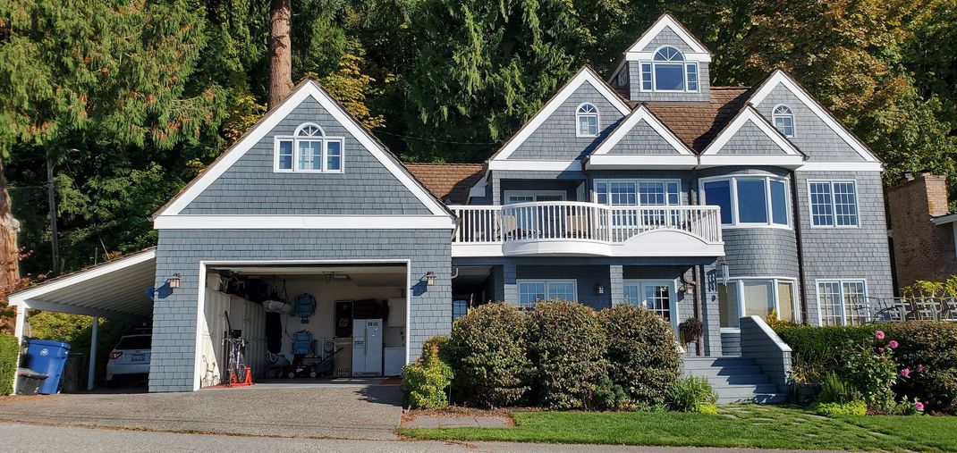 Large gray house with blue trim, garage, and front steps surrounded by trees and shrubs