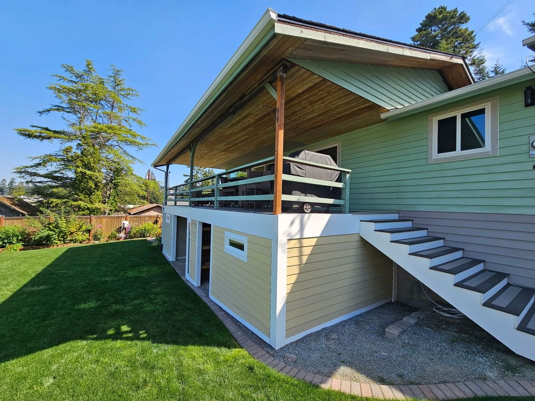 Two-story house with green siding, covered deck, and exterior staircase beside a manicured lawn.