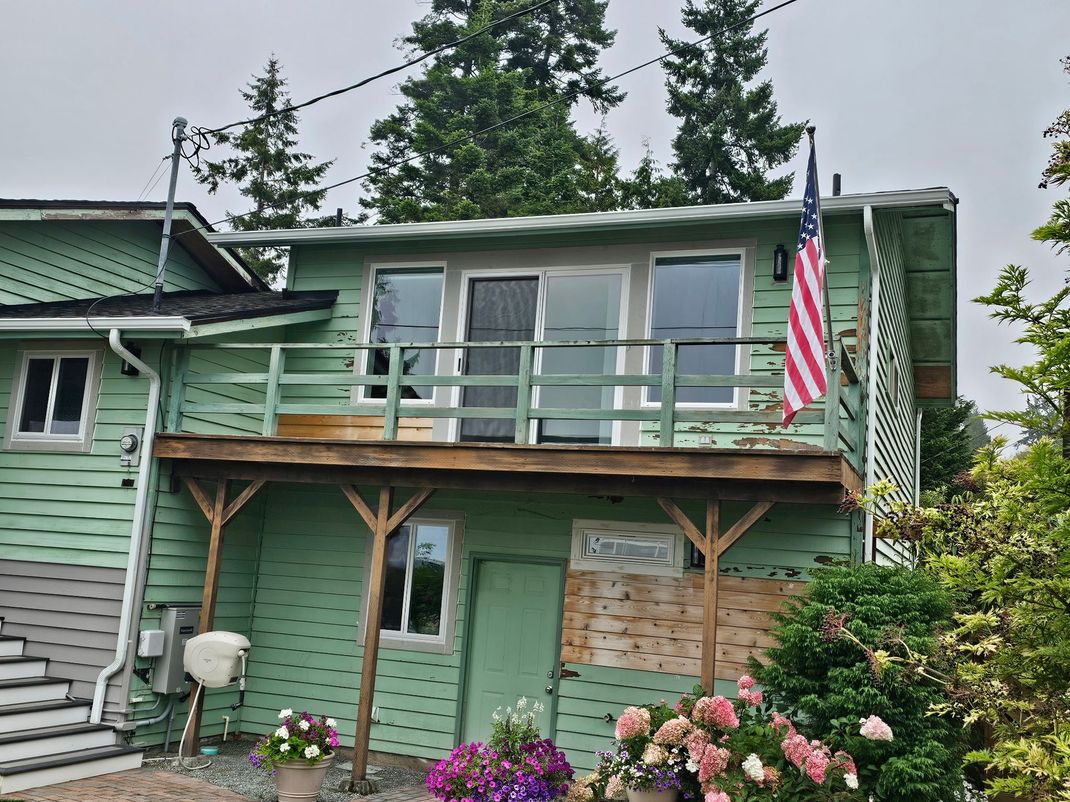 Green two-story house with porch, American flag, and pink flowers in front yard.