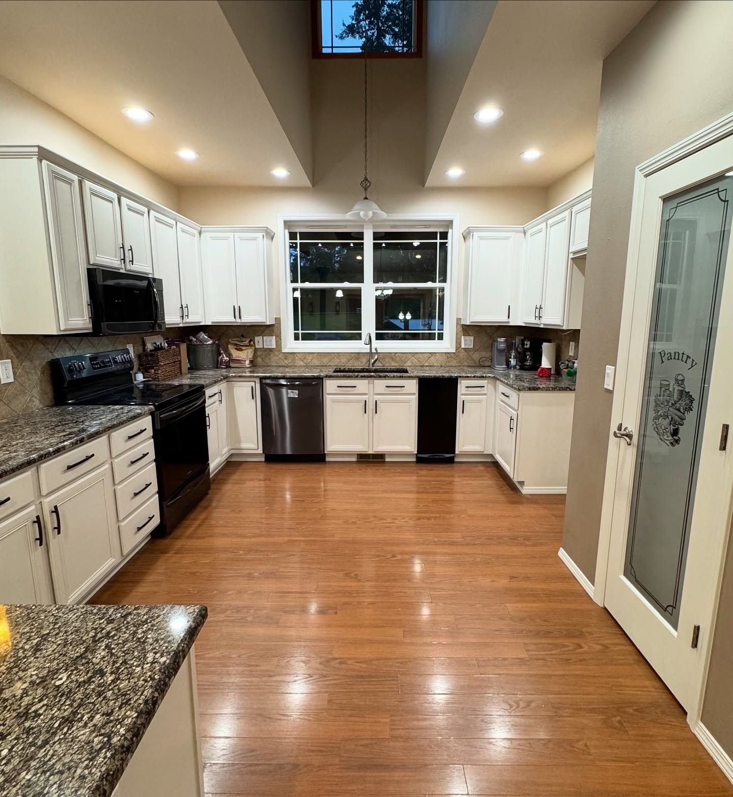 Bright kitchen with white cabinets, stainless appliances, wood floors, and a central window.