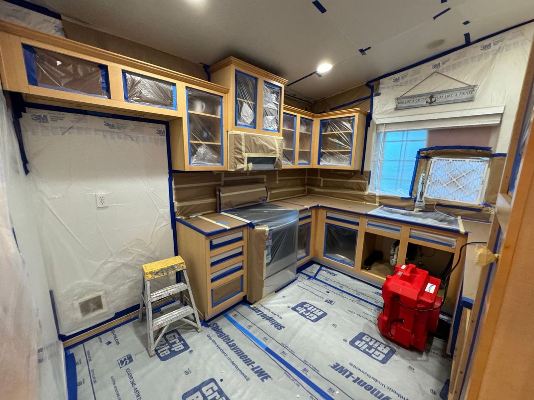 Unfinished kitchen with wood cabinets, blue tape-covered surfaces, ladder, and a window on the right.