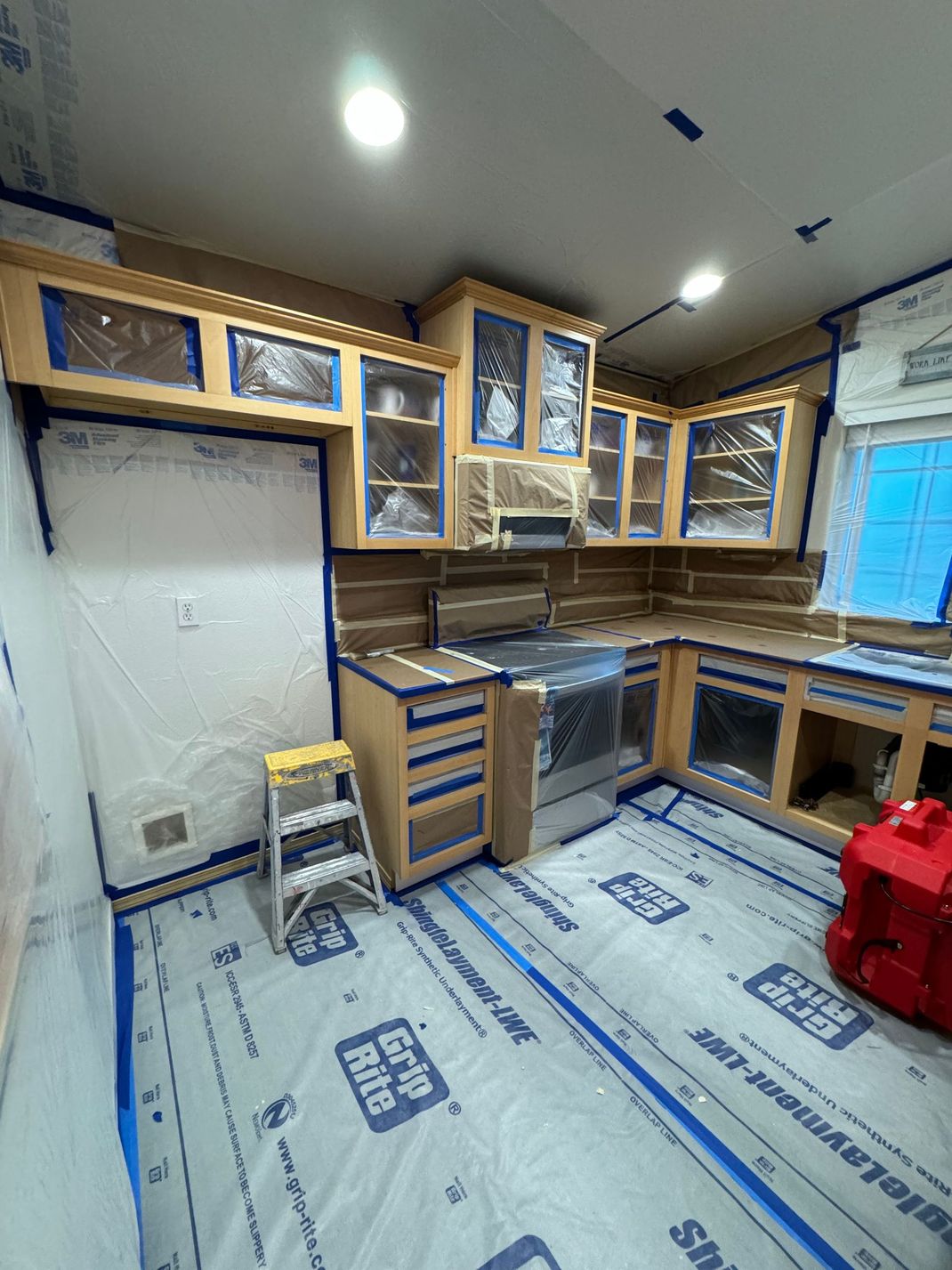 Unfinished kitchen with wood cabinets, countertops, covered floor, and blue painter’s tape on walls and ceiling.