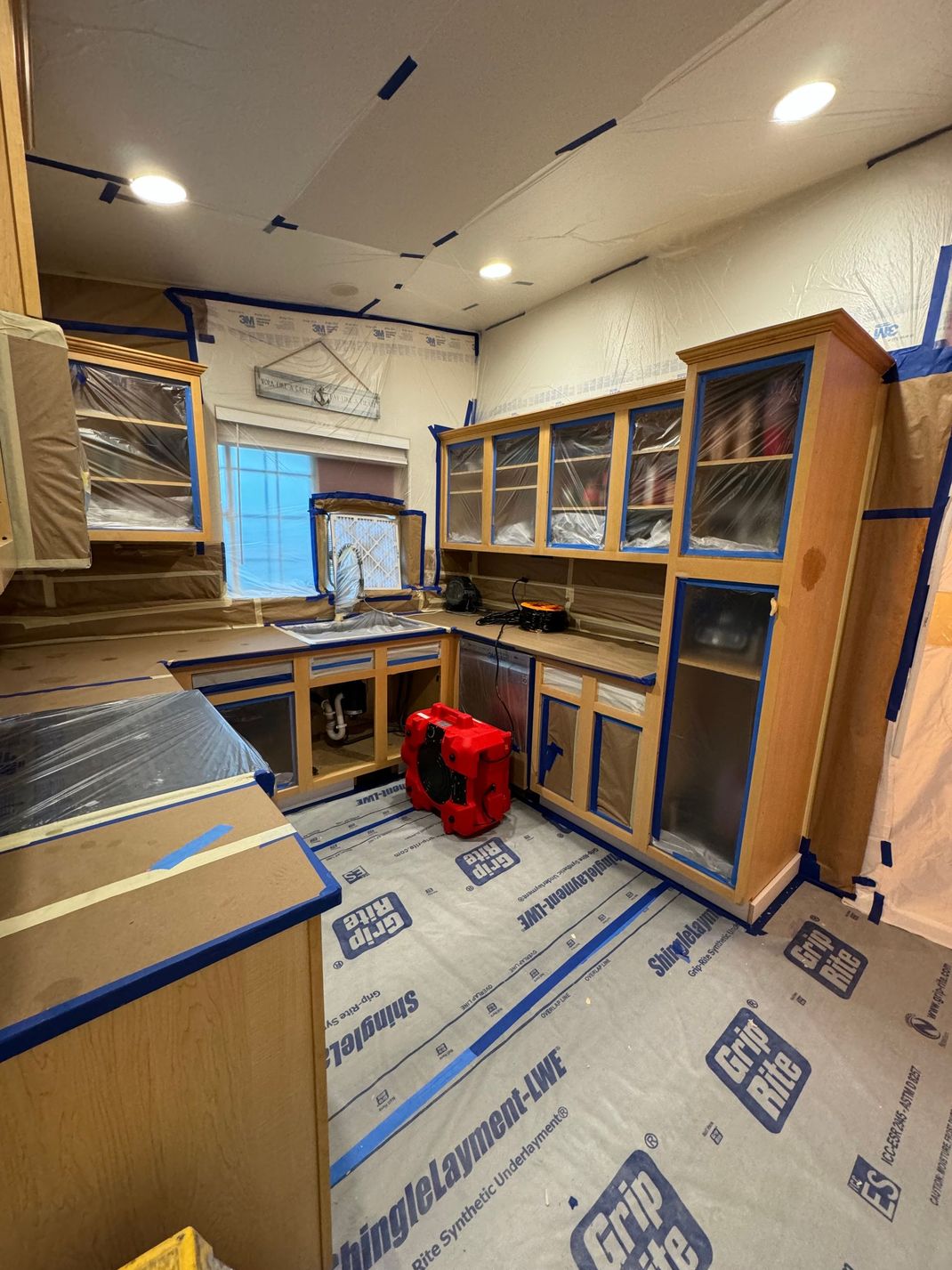 Kitchen under renovation with new cabinets, exposed flooring, and a red vacuum near the window