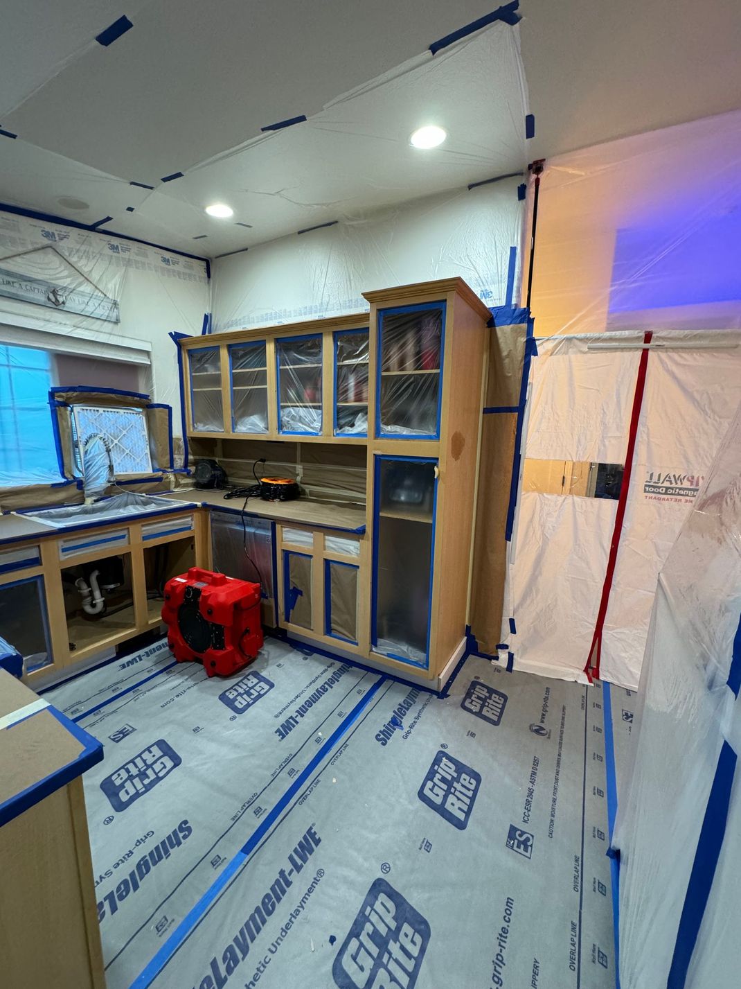 Kitchen under renovation with covered floors, cabinets, and a red fan on the floor