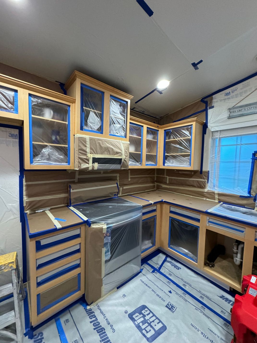 Compact kitchen under renovation with gray cabinets, stainless stove, blue-taped windows, and protective floor covering