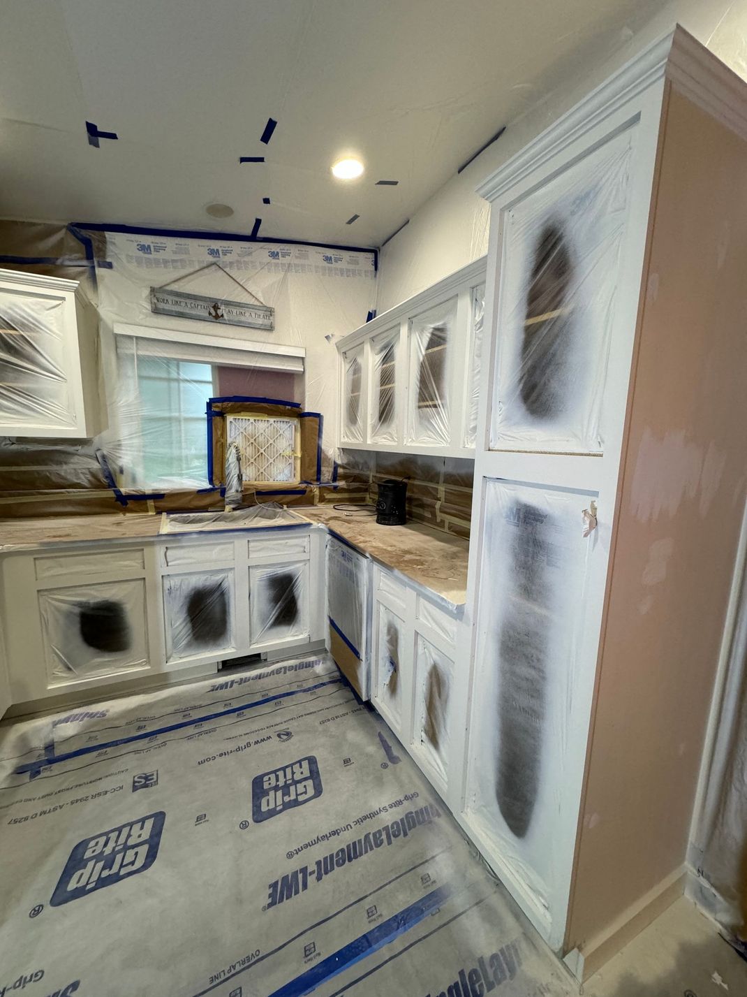 Small bright kitchen with white cabinets, window, countertop appliances, and protective floor covering during renovation