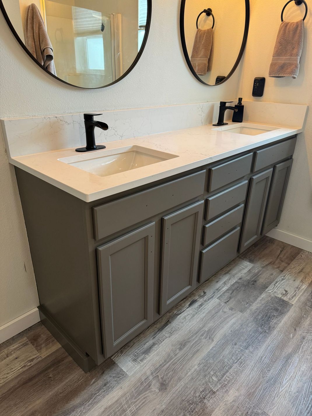 Modern bathroom vanity with double sinks, matte black faucets, oval mirrors, and gray cabinets on wood-look flooring