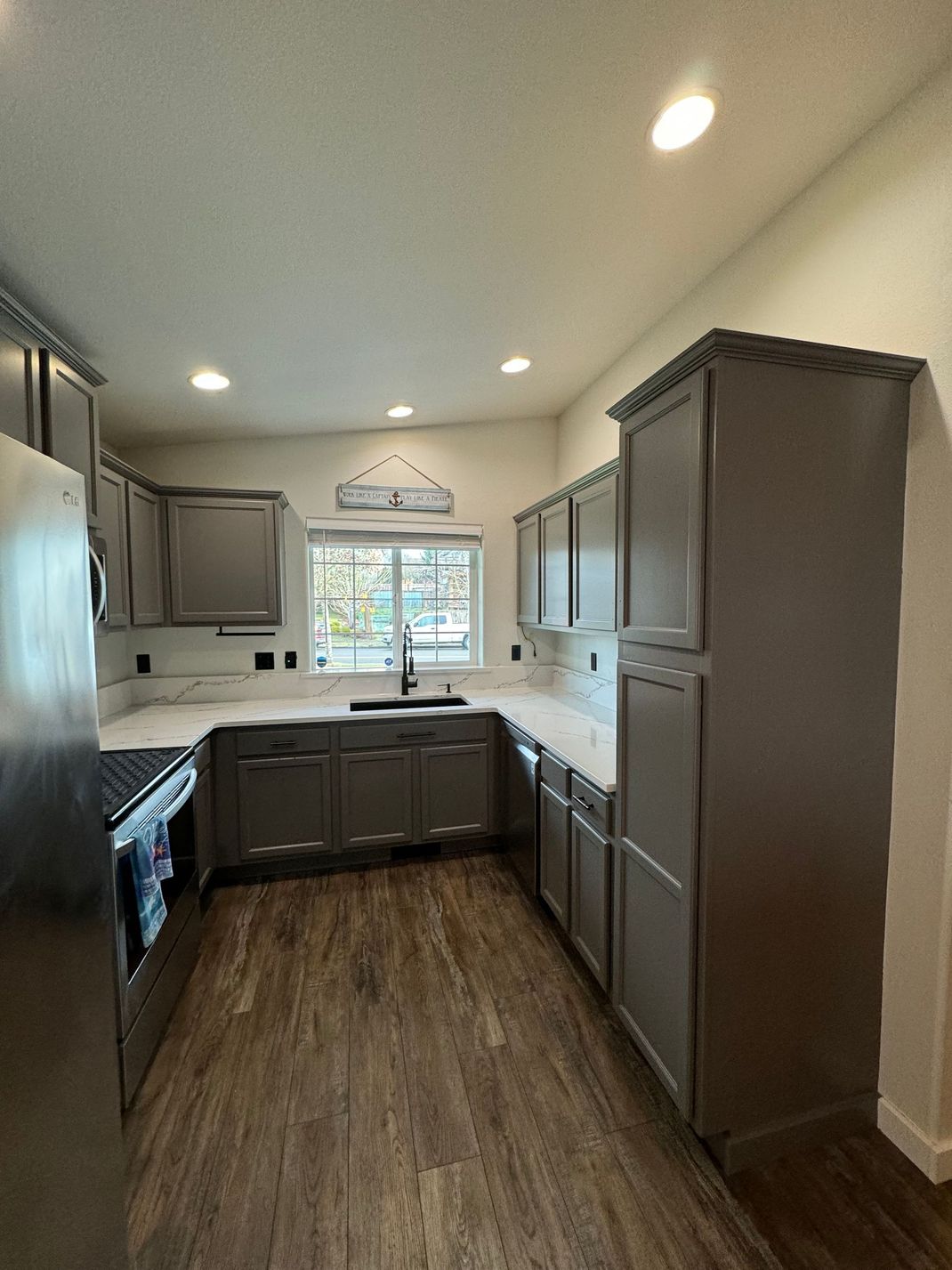 Small modern kitchen with dark cabinets, stainless stove, and wood-look floors, lit by recessed ceiling lights.
