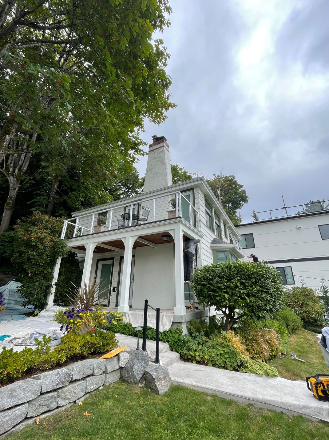 White Victorian-style house with porch and chimney, surrounded by lush gardens and trees under a cloudy sky