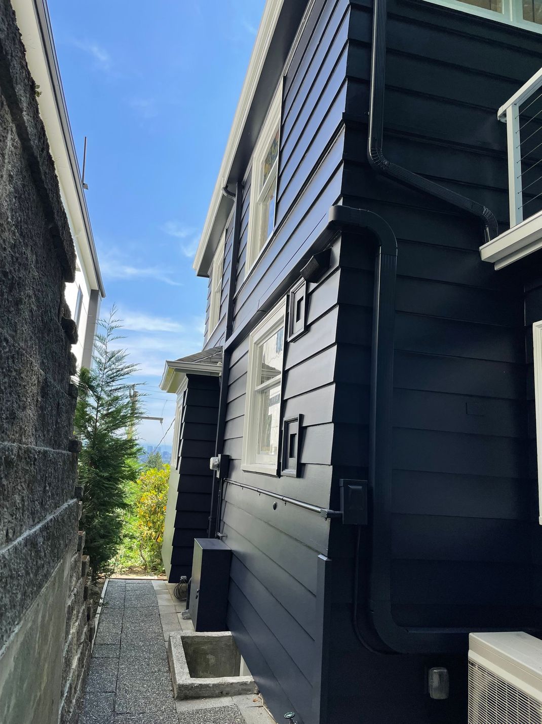 Narrow outdoor alley between dark-clad buildings with blue sky overhead and a small patch of greenery ahead