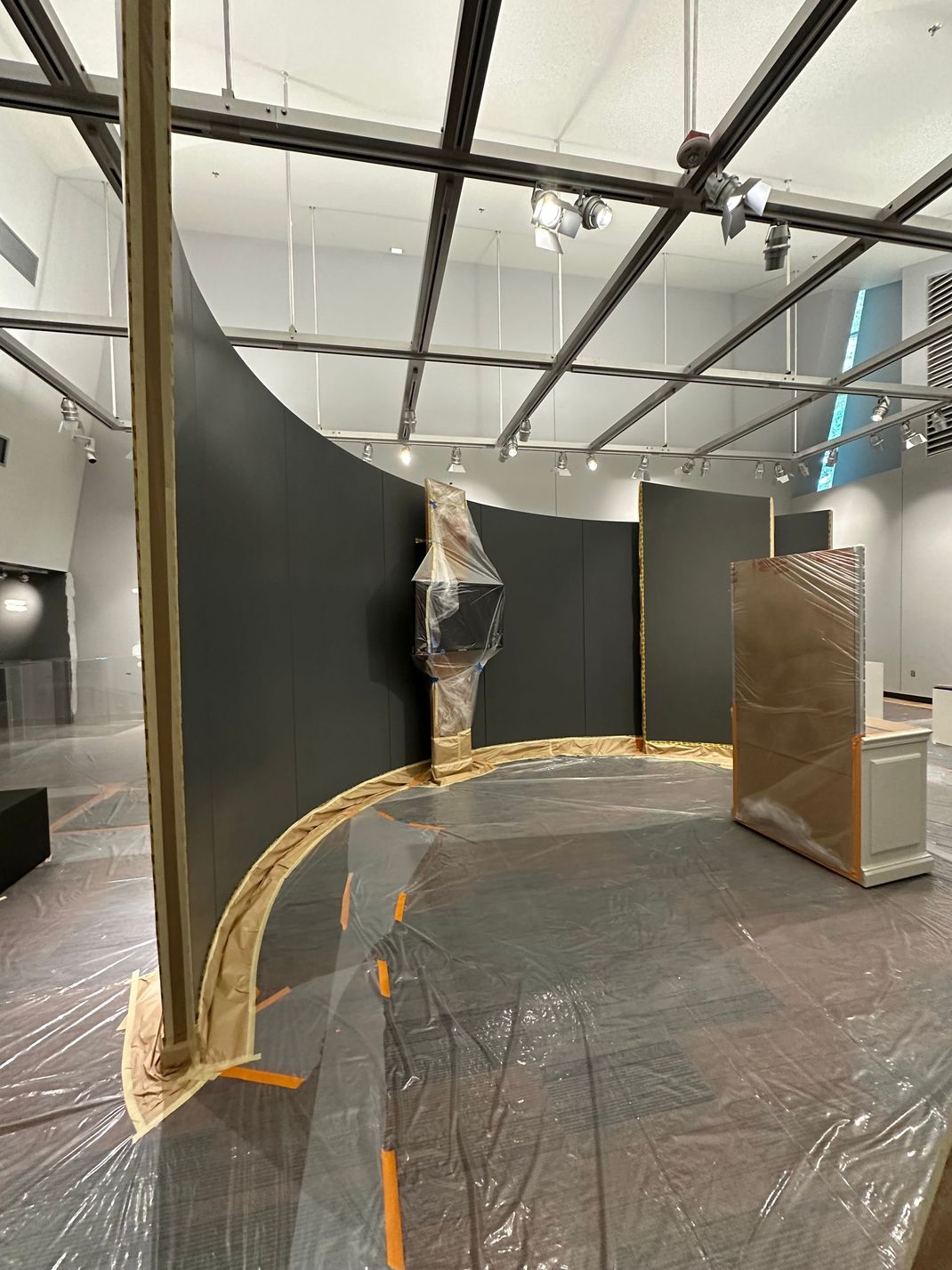 Curved black walls and plastic-covered floor in an art installation space, with a central hanging figure.