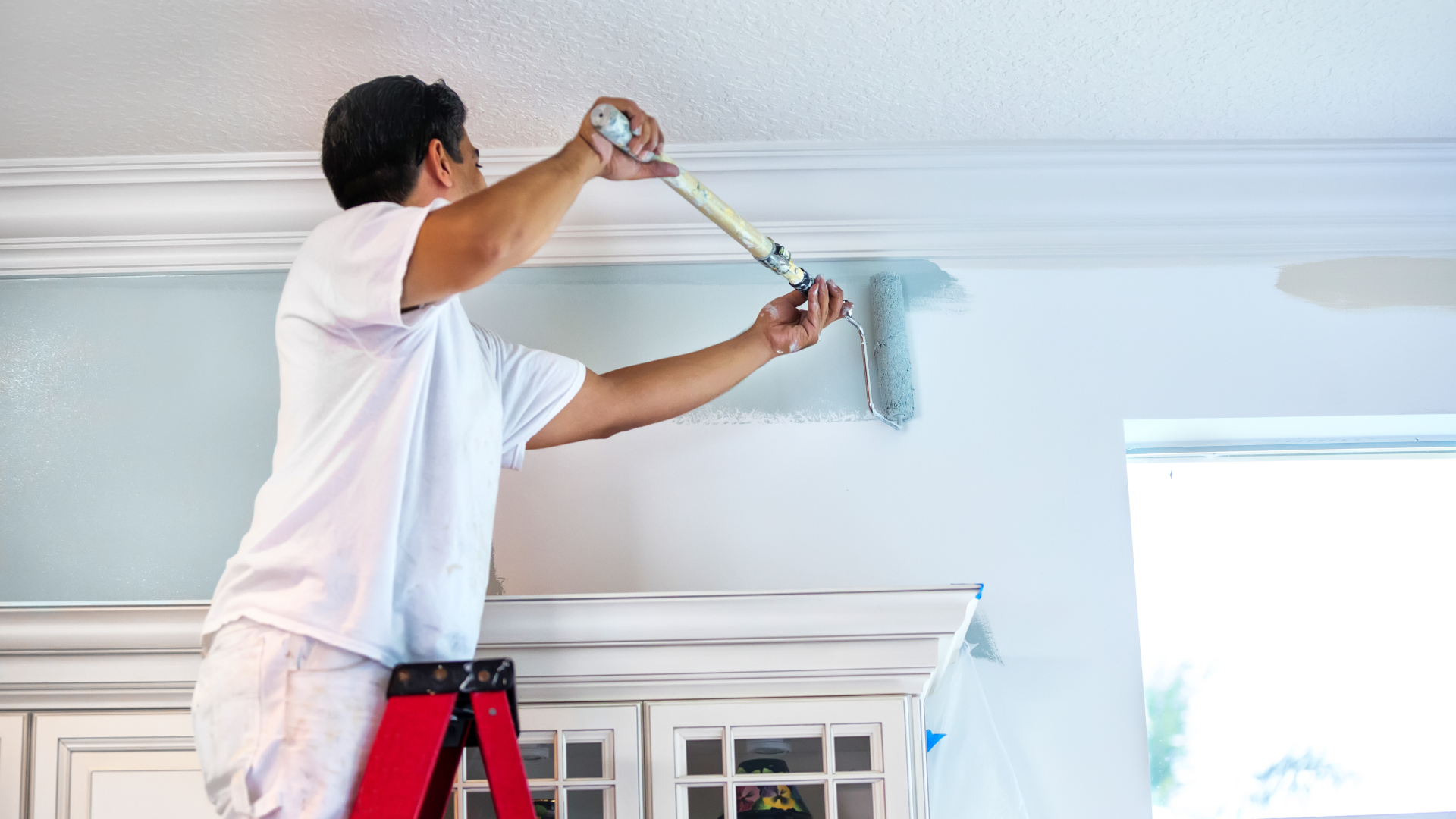 Person painting a light blue ceiling trim with a roller in a bright room