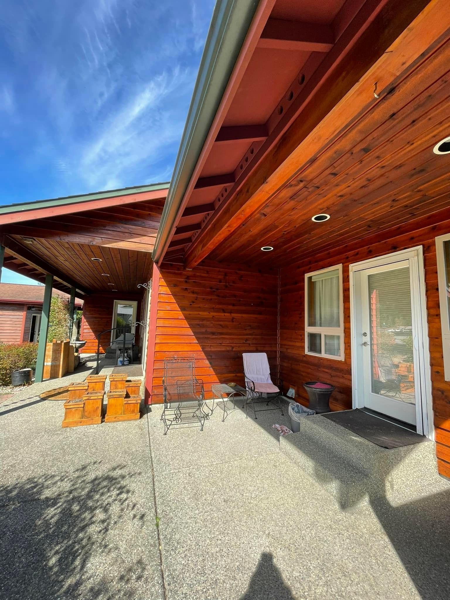 A shaded patio with wooden walls and ceiling, outdoor chairs, and a sliding glass door under a wide roof overhang.