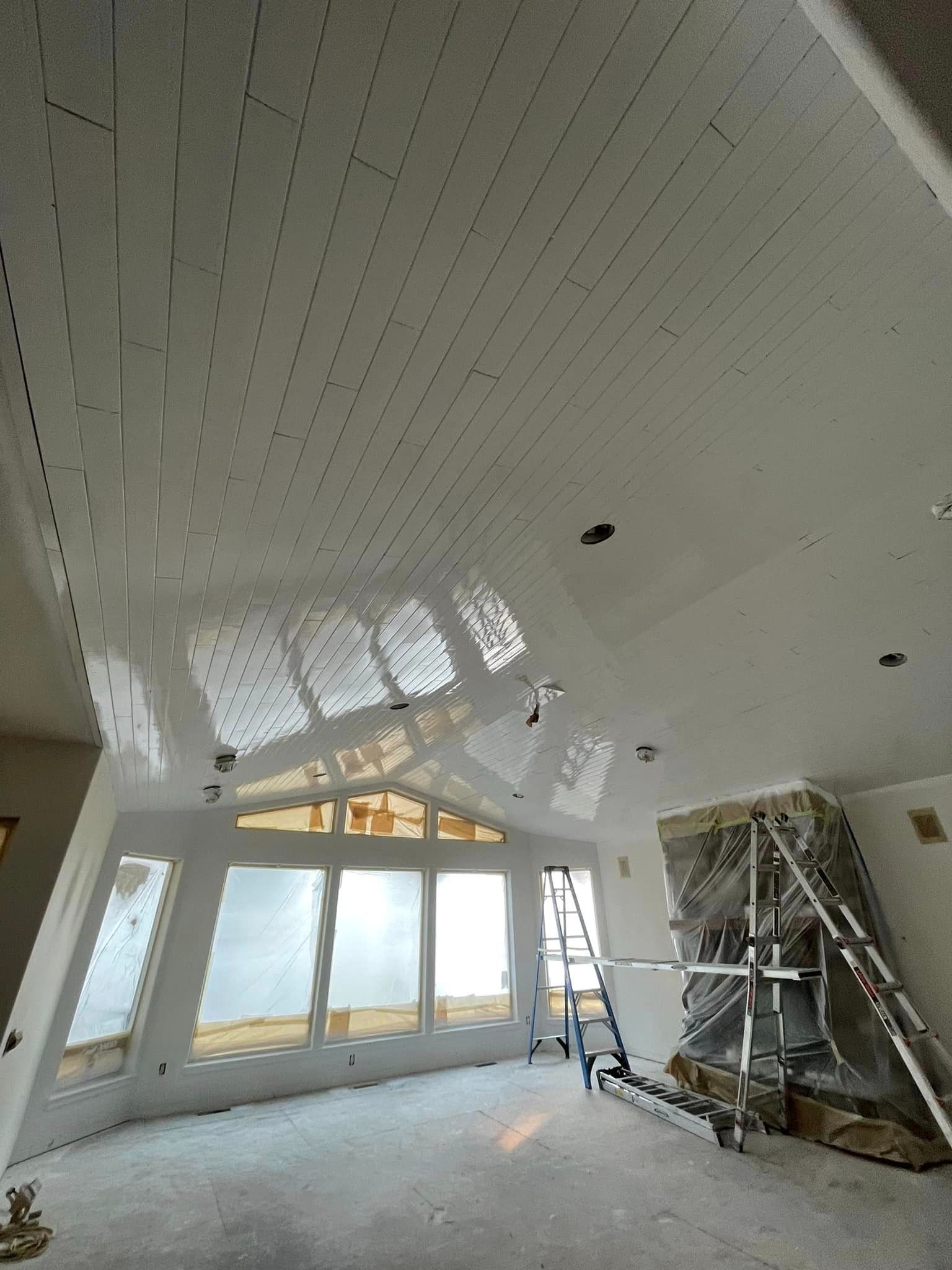 A room under construction with a high, white paneled ceiling, large windows, and a tall metal ladder by a fireplace.