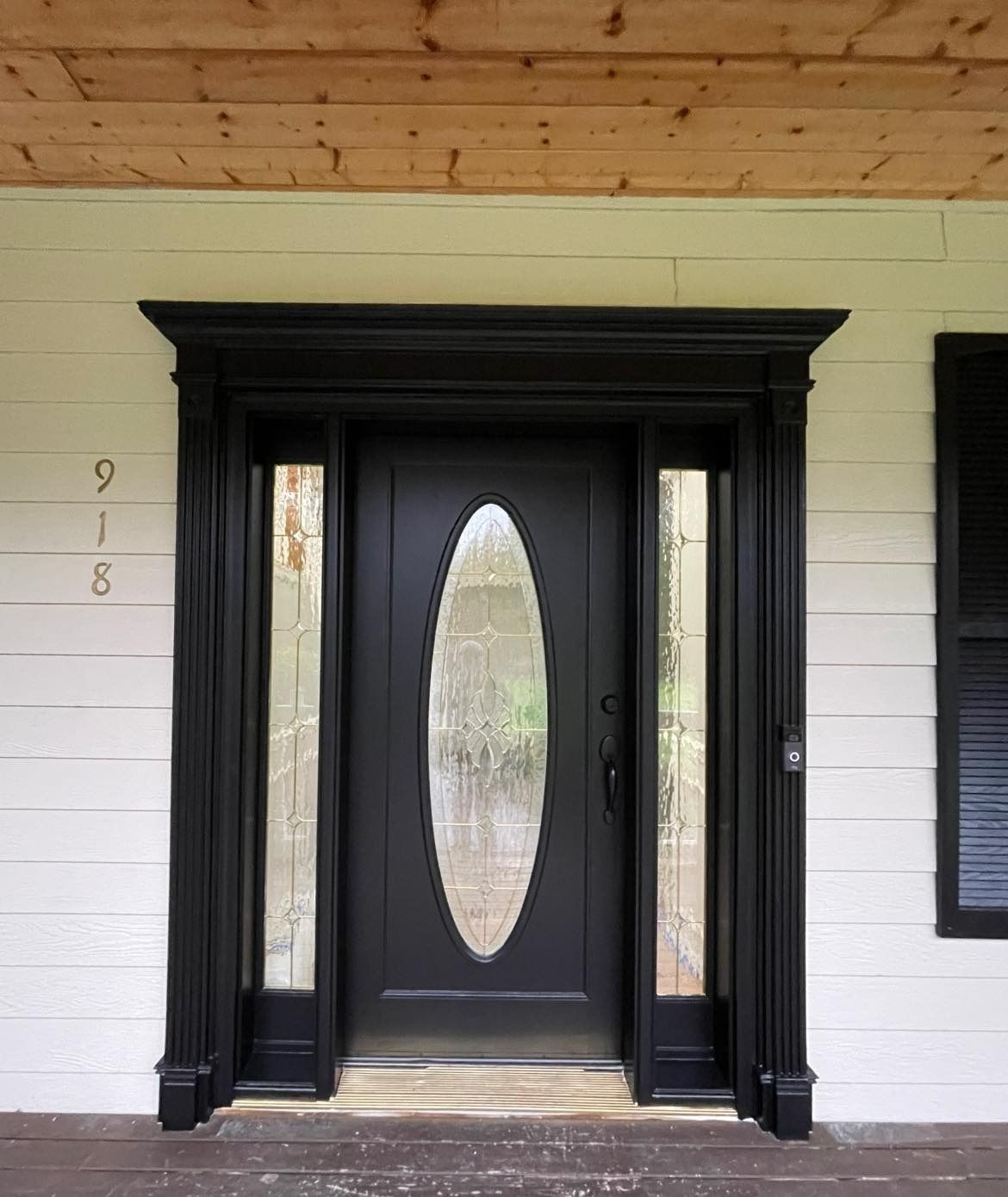 A black front door with an oval glass insert, flanked by vertical sidelights, set in a white-walled house with wood ceiling.
