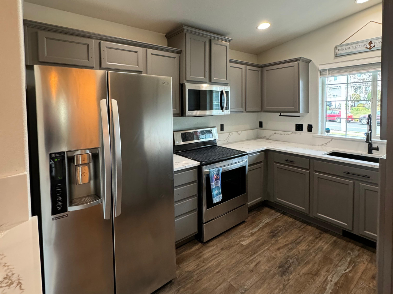 A modern kitchen with stainless steel appliances, gray cabinets, white countertops, and wood-look flooring.