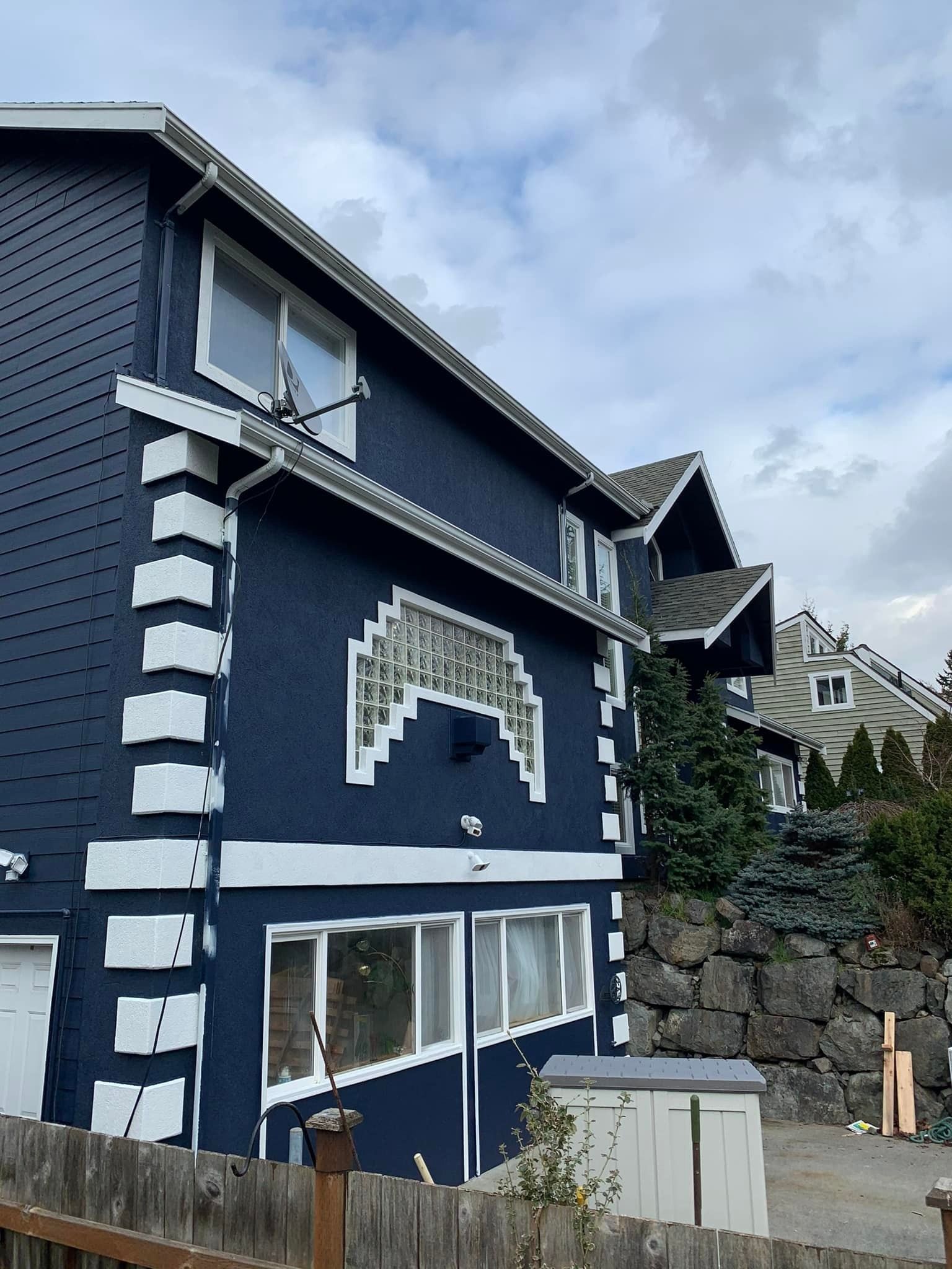 A dark blue house with white decorative corner stones and trim, set against a stone retaining wall under a cloudy sky.