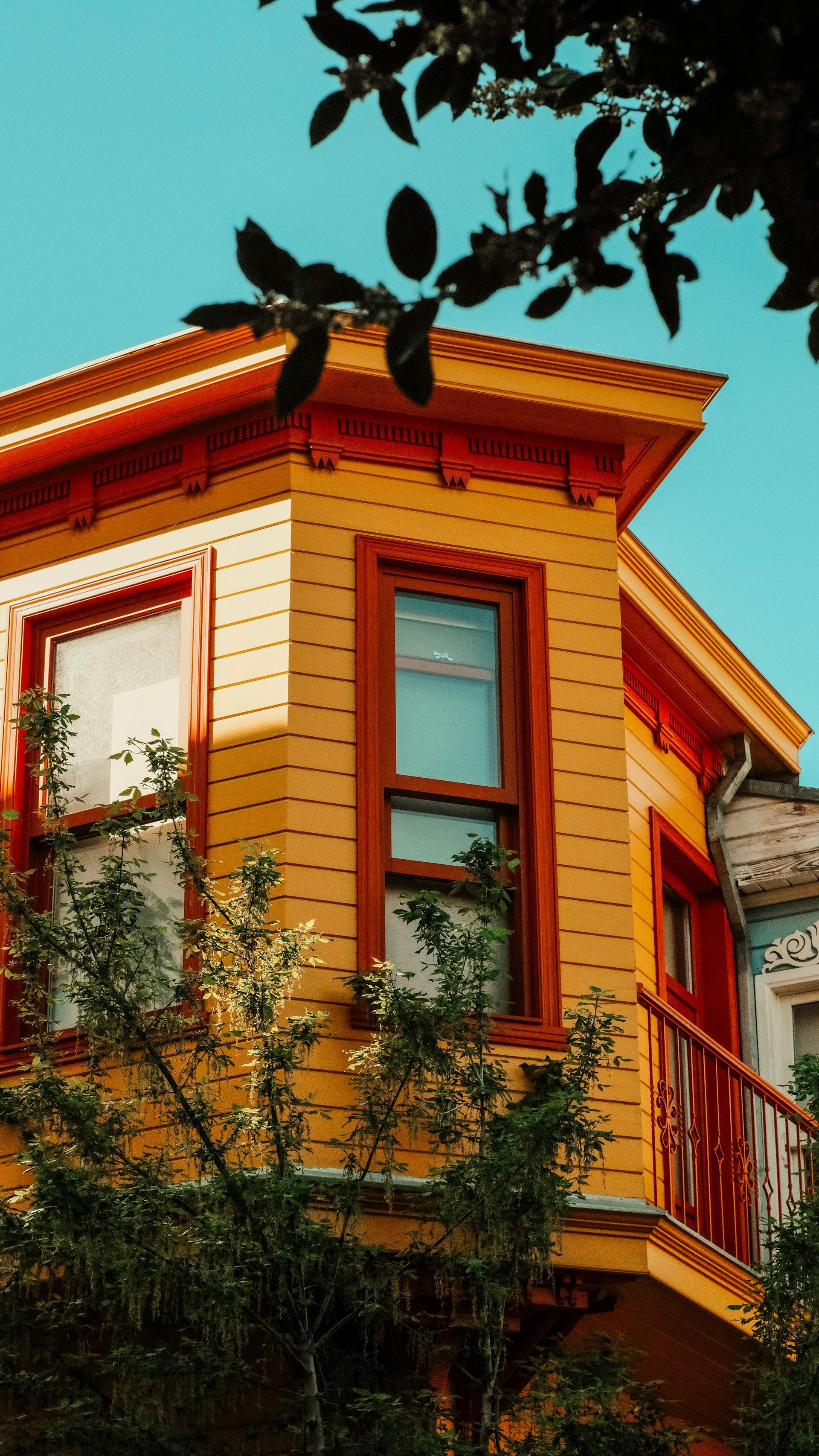 Orange-trimmed bay window on a yellow house with leafy branches against a bright blue sky