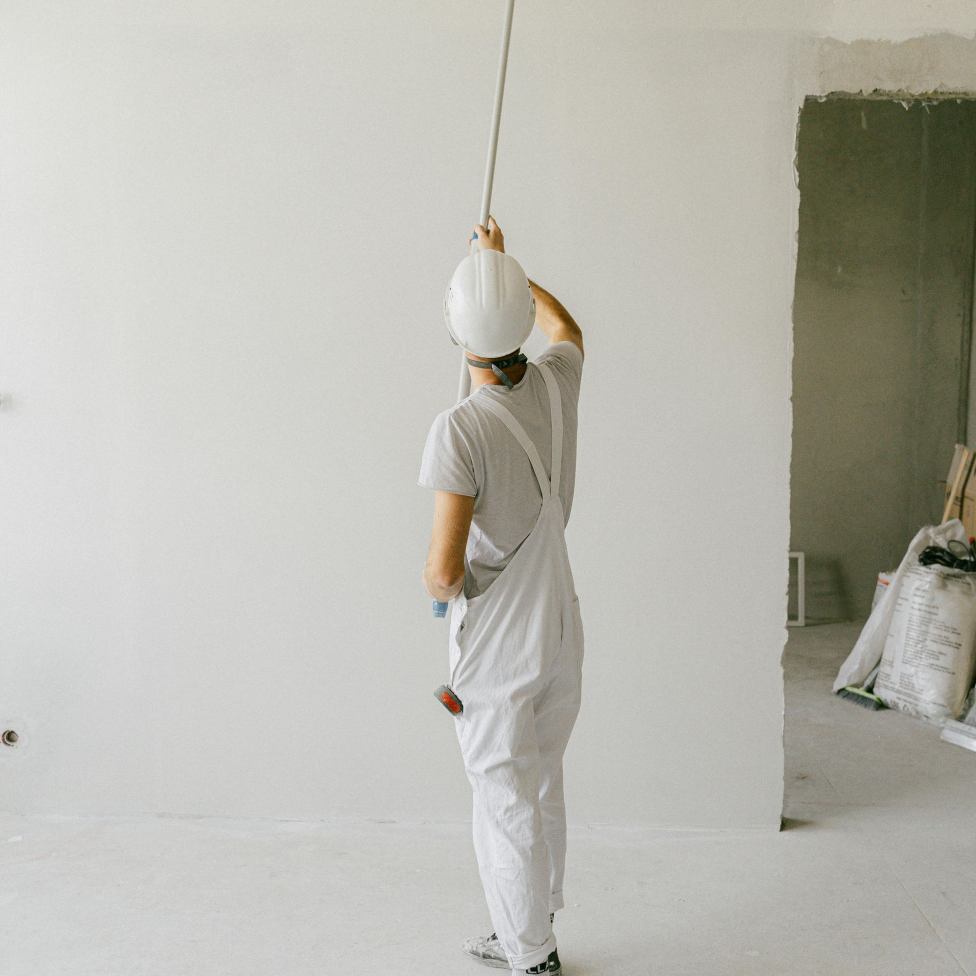 Person in white overalls and hard hat painting a white wall in a room under renovation.