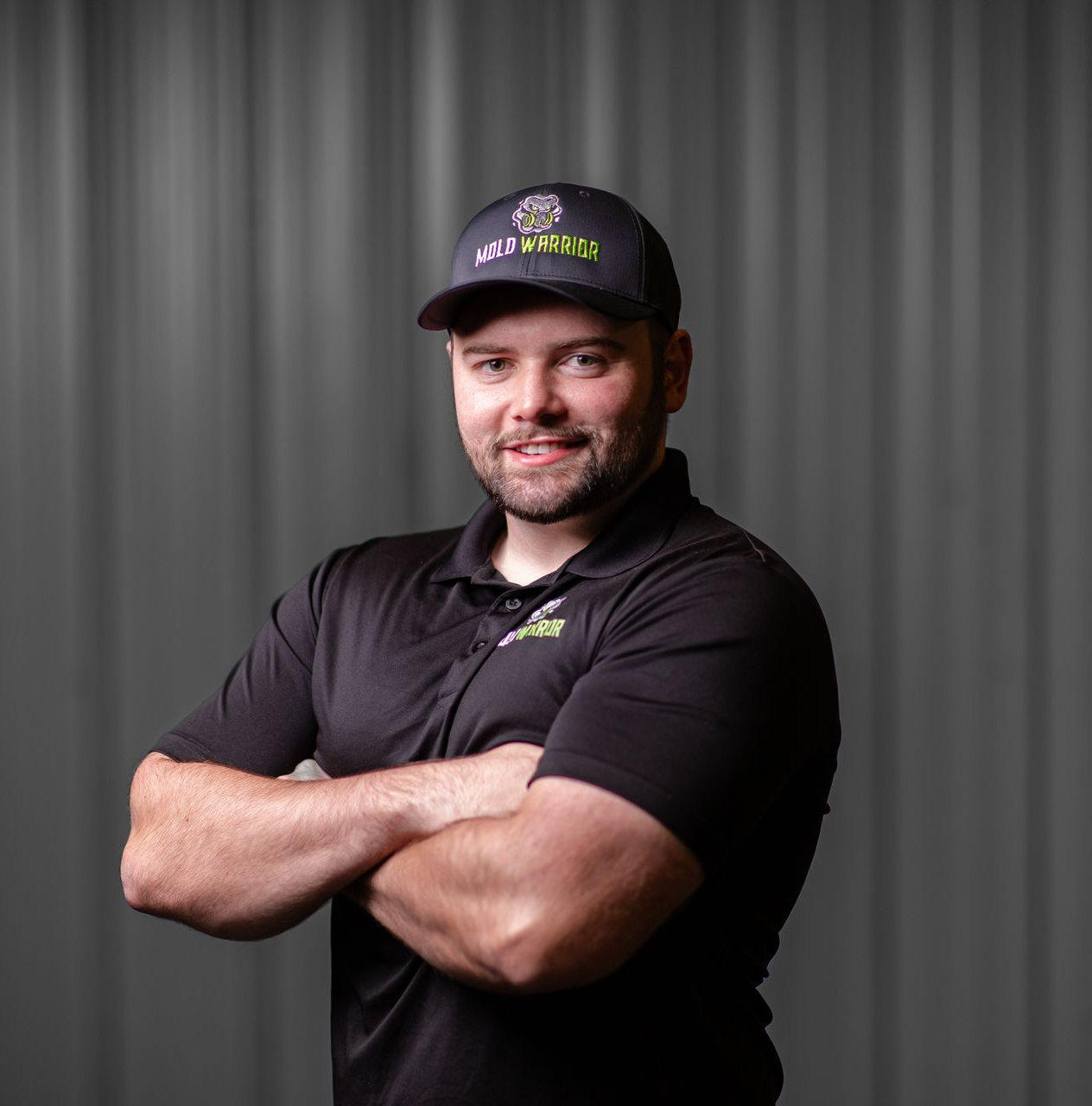 Man in black cap and shirt, arms crossed, smiles, stands in front of gray background.