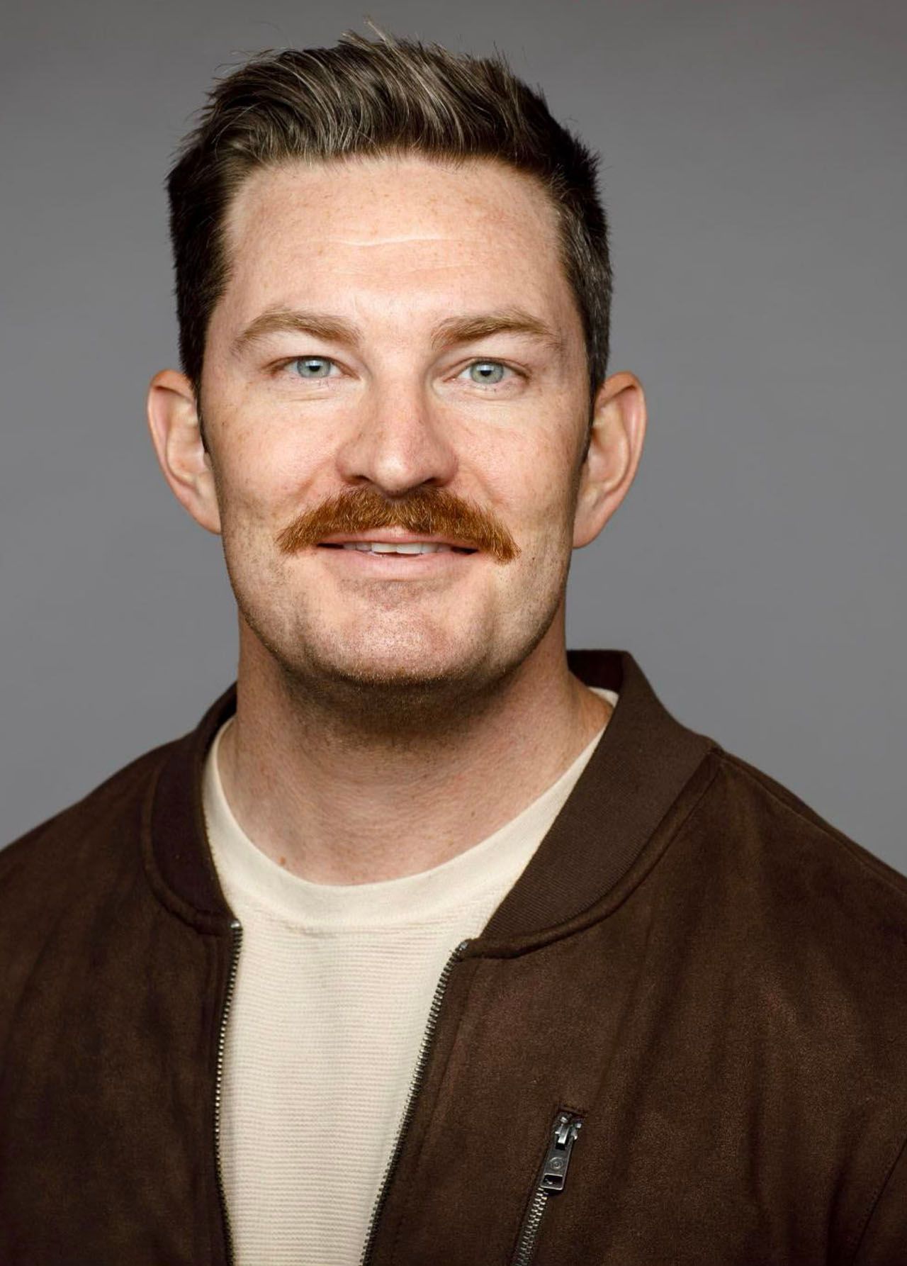 Man with brown hair and mustache, wearing a brown bomber jacket, smiling.