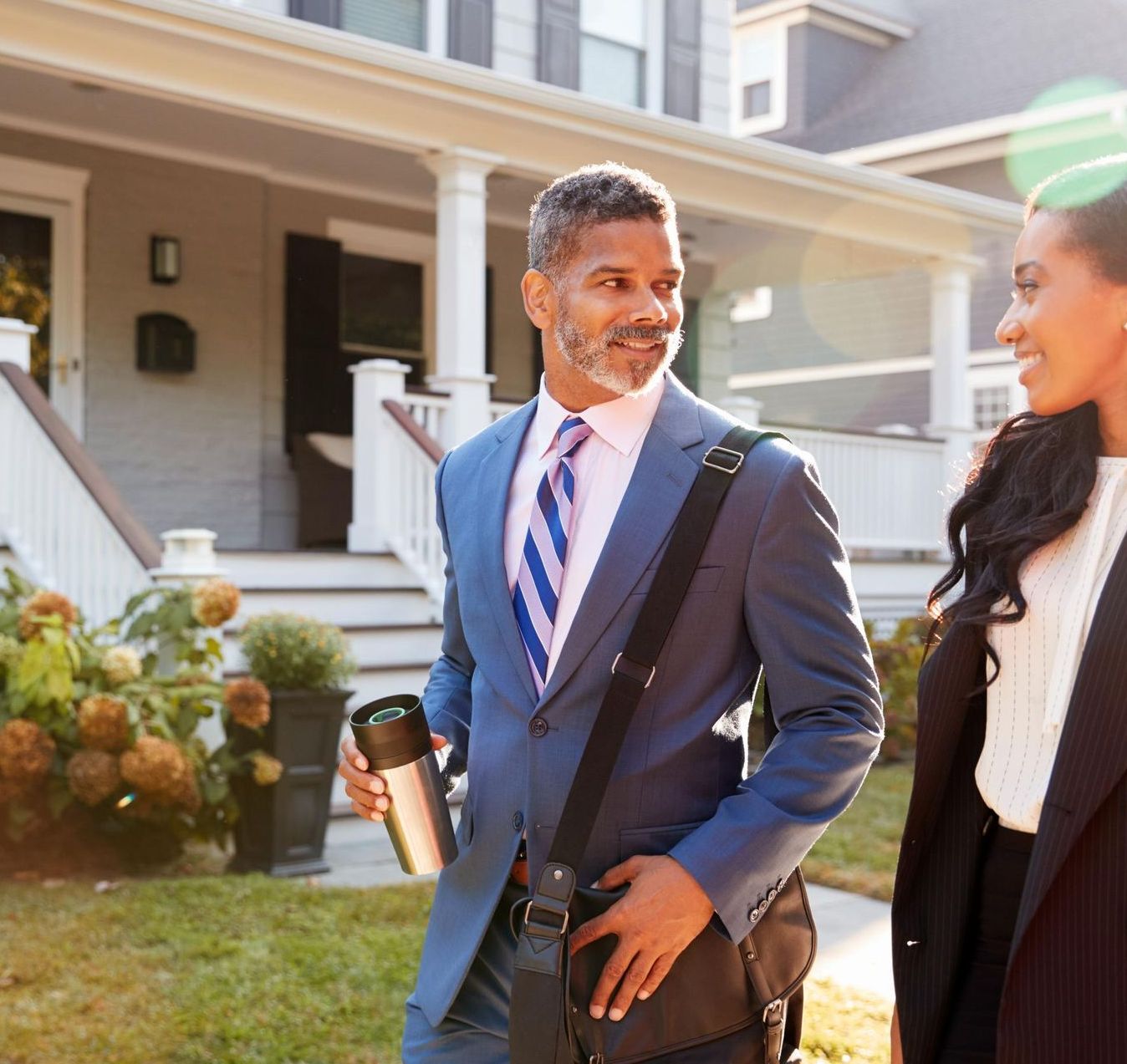 A man in a suit and a woman walk on a sidewalk in front of a house, smiling, talking.