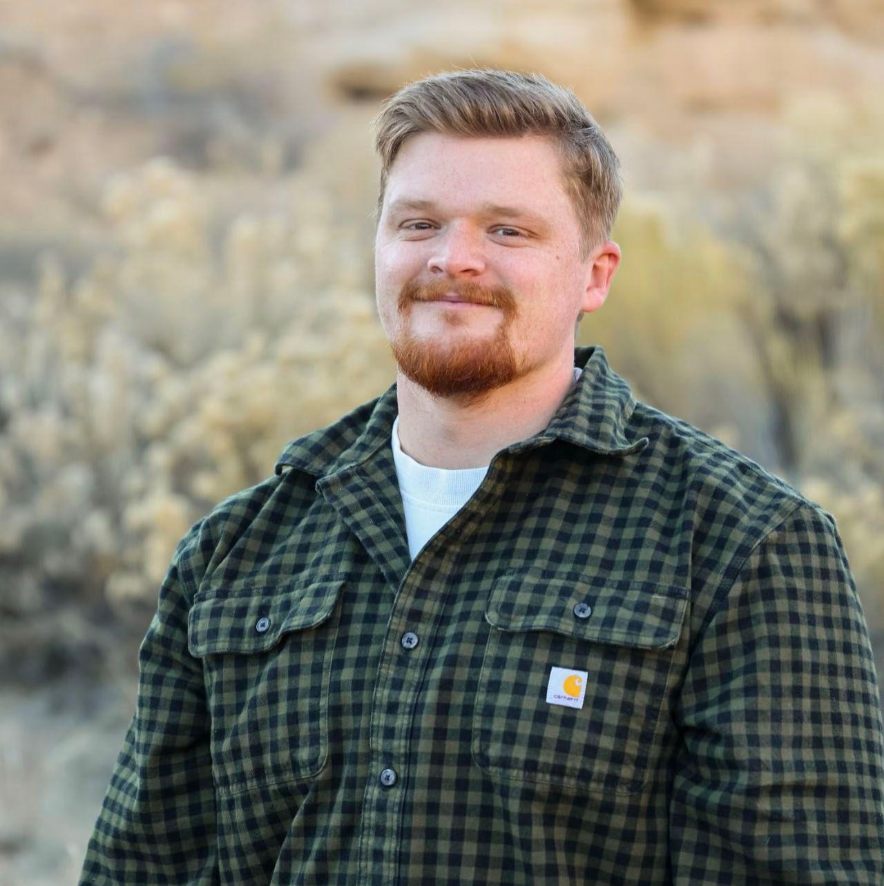 Man with red beard smiles, wearing a green plaid shirt and white shirt outdoors.