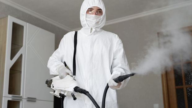 Person in white hazmat suit sanitizing room with a steam cleaner.
