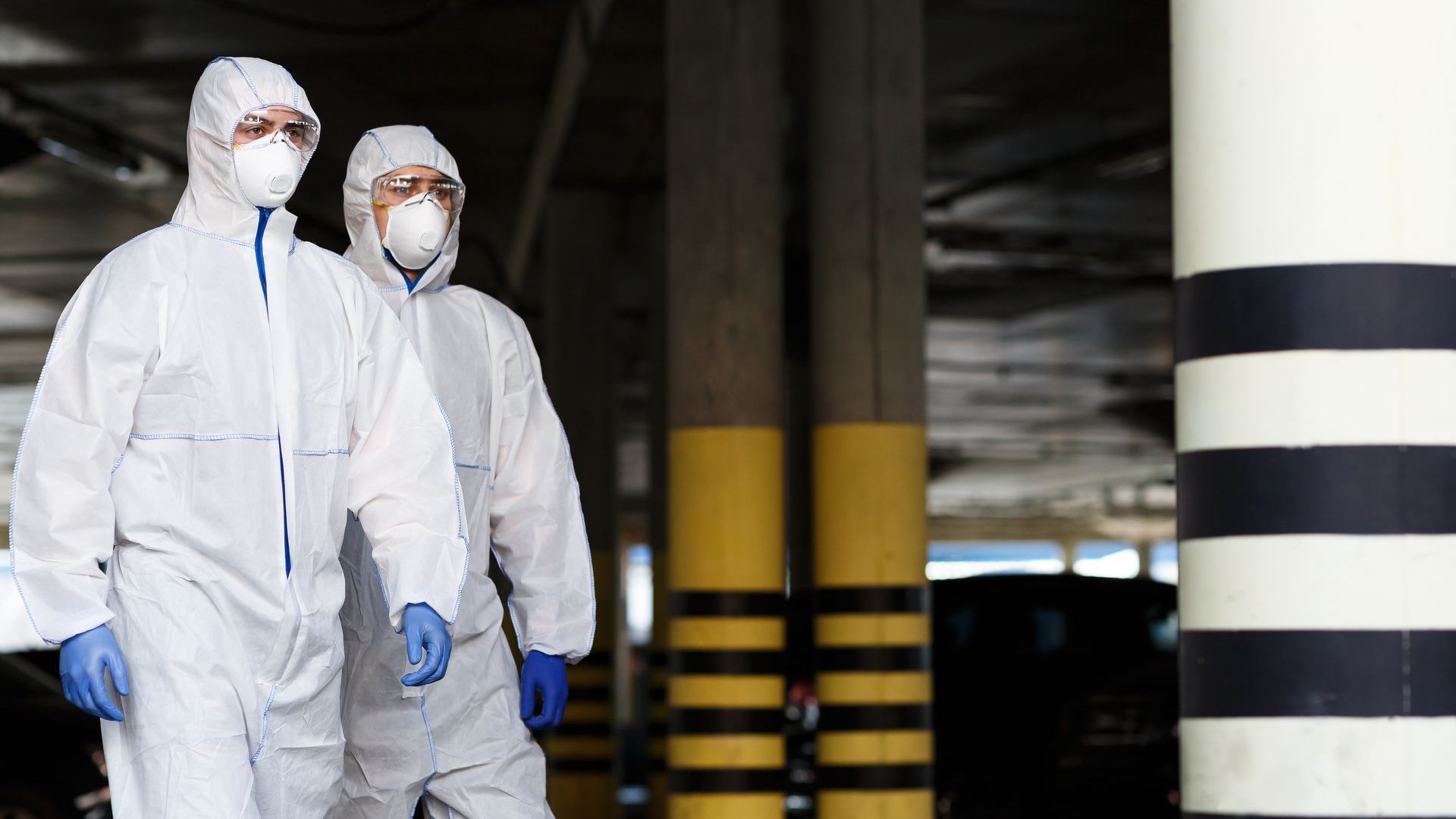 Two people in white hazmat suits walk through a parking garage.