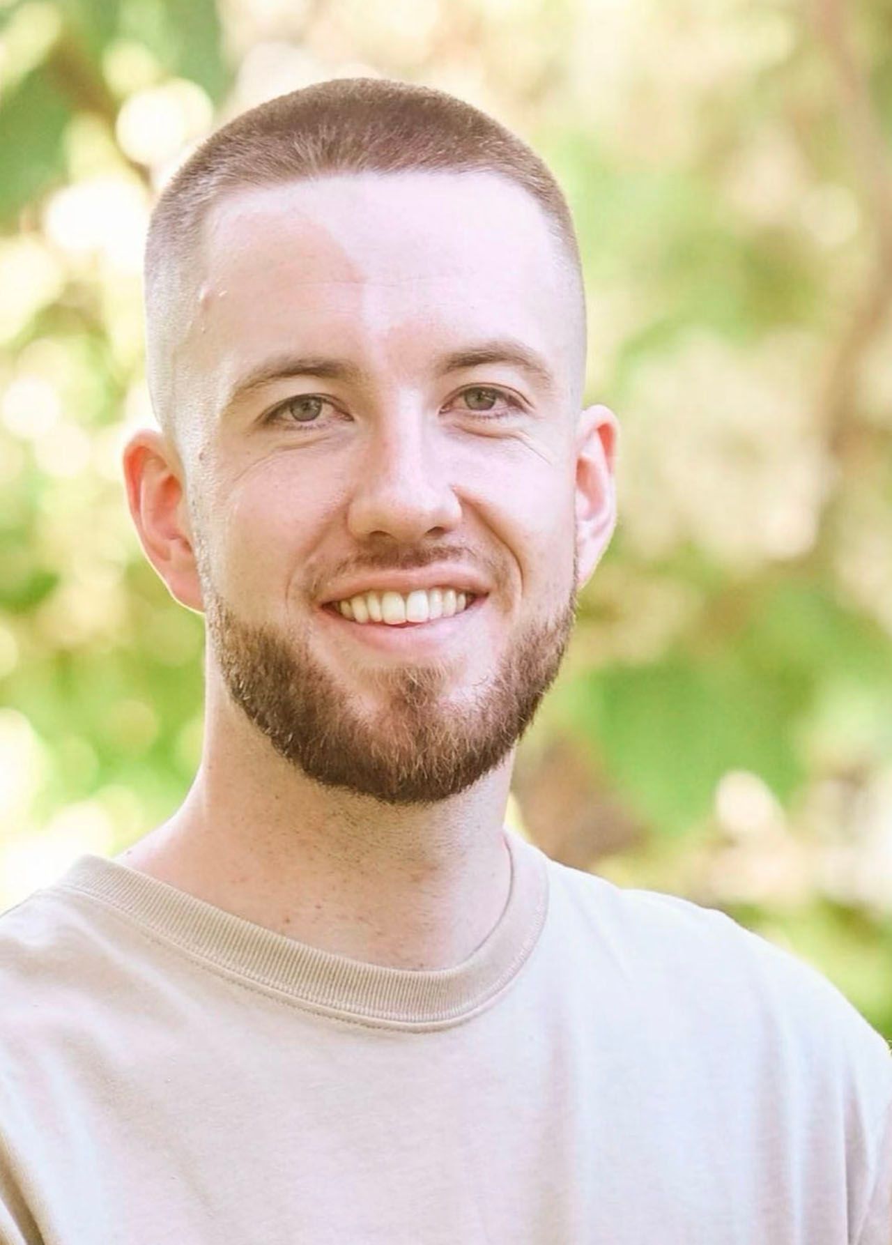 Young man with a short haircut and beard smiling, wearing a beige shirt, outdoors with green background.