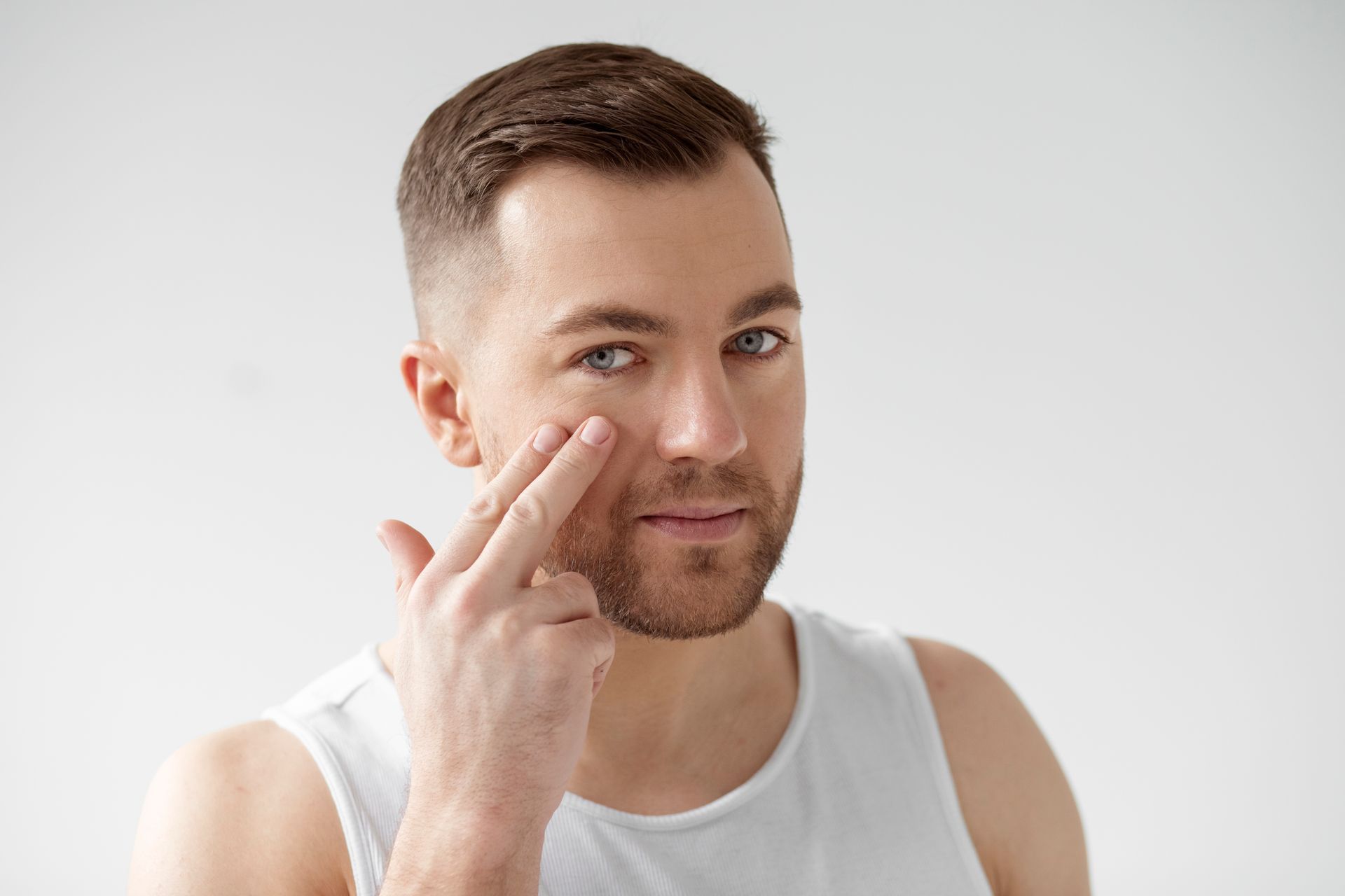 A man in a white tank top is applying lotion to his face.
