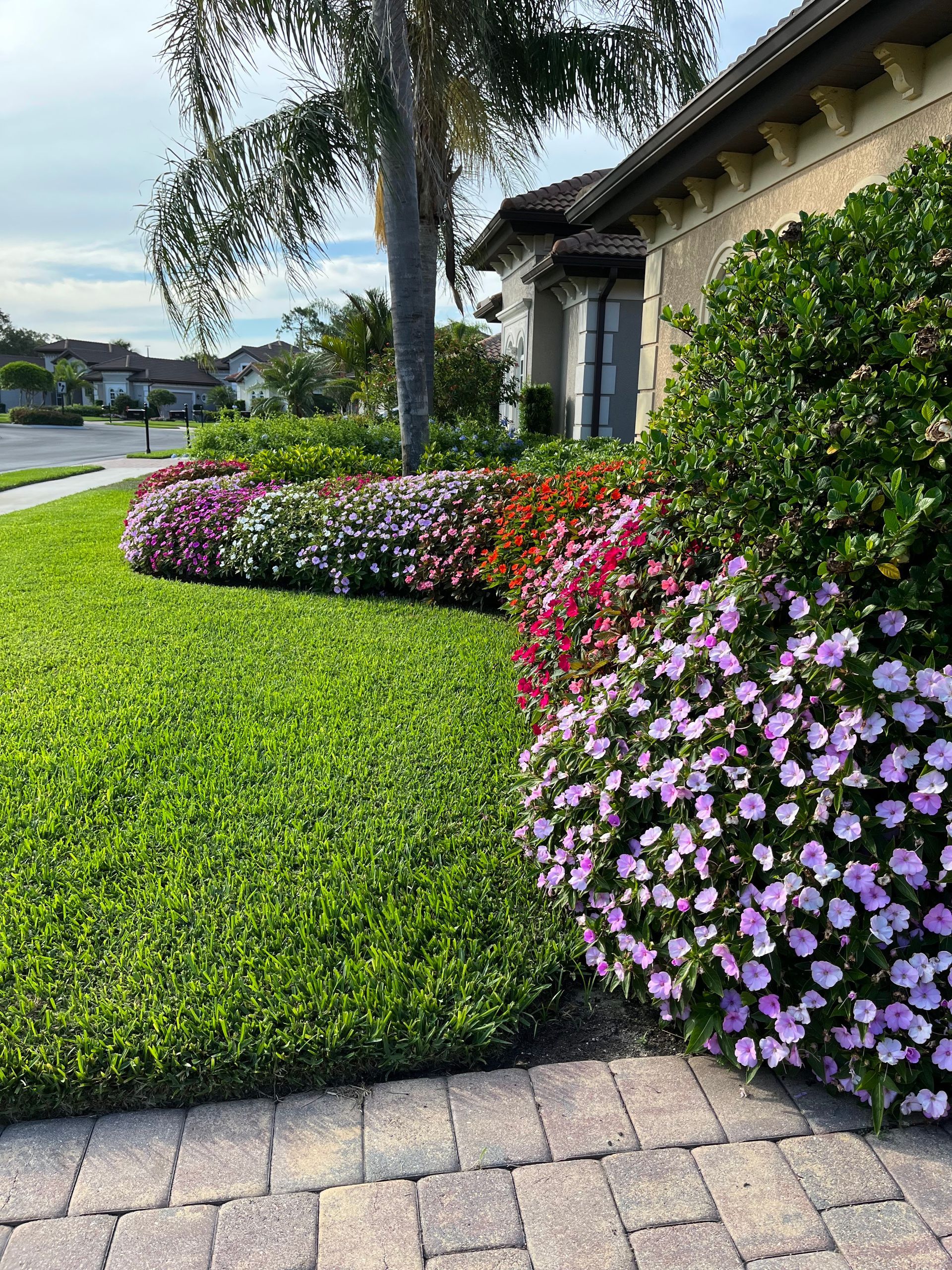 Lush green lawn borders a vibrant flower bed with pink, red, and white blooms in front of a house.