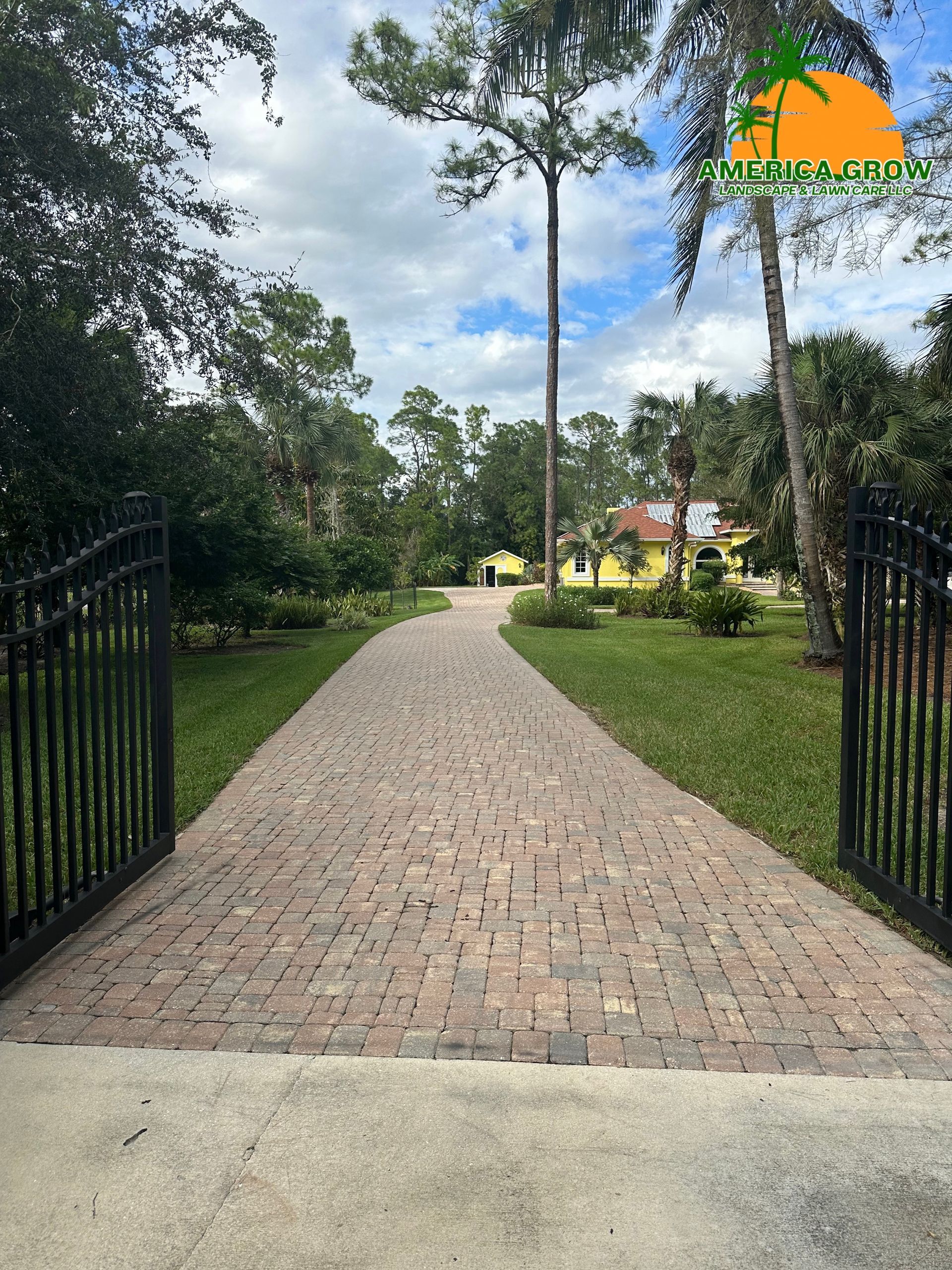 Brick driveway leading to a yellow house, flanked by green grass and trees, through open black gates.