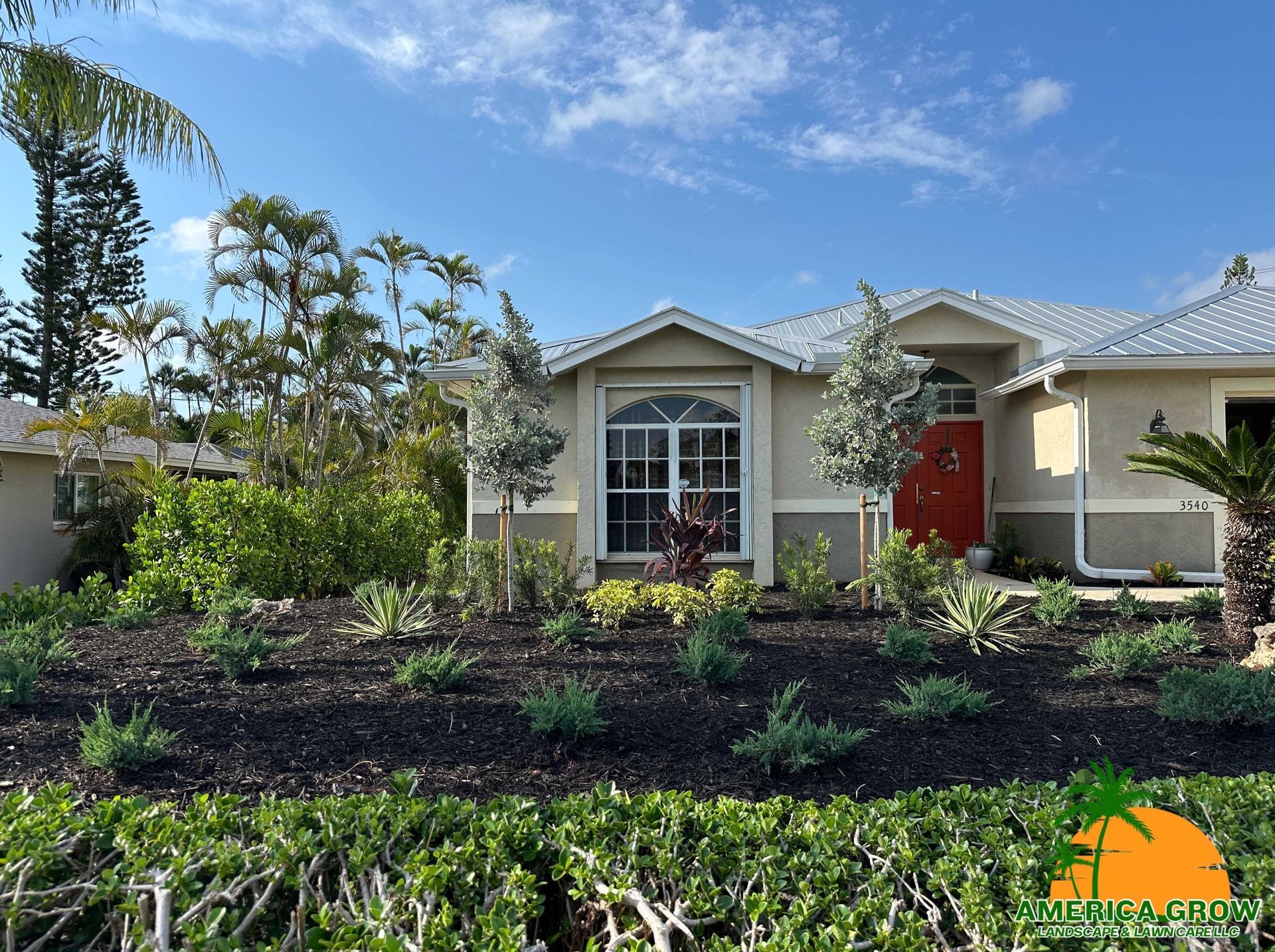 Tan house with a red door, lush landscaping, and a blue sky.