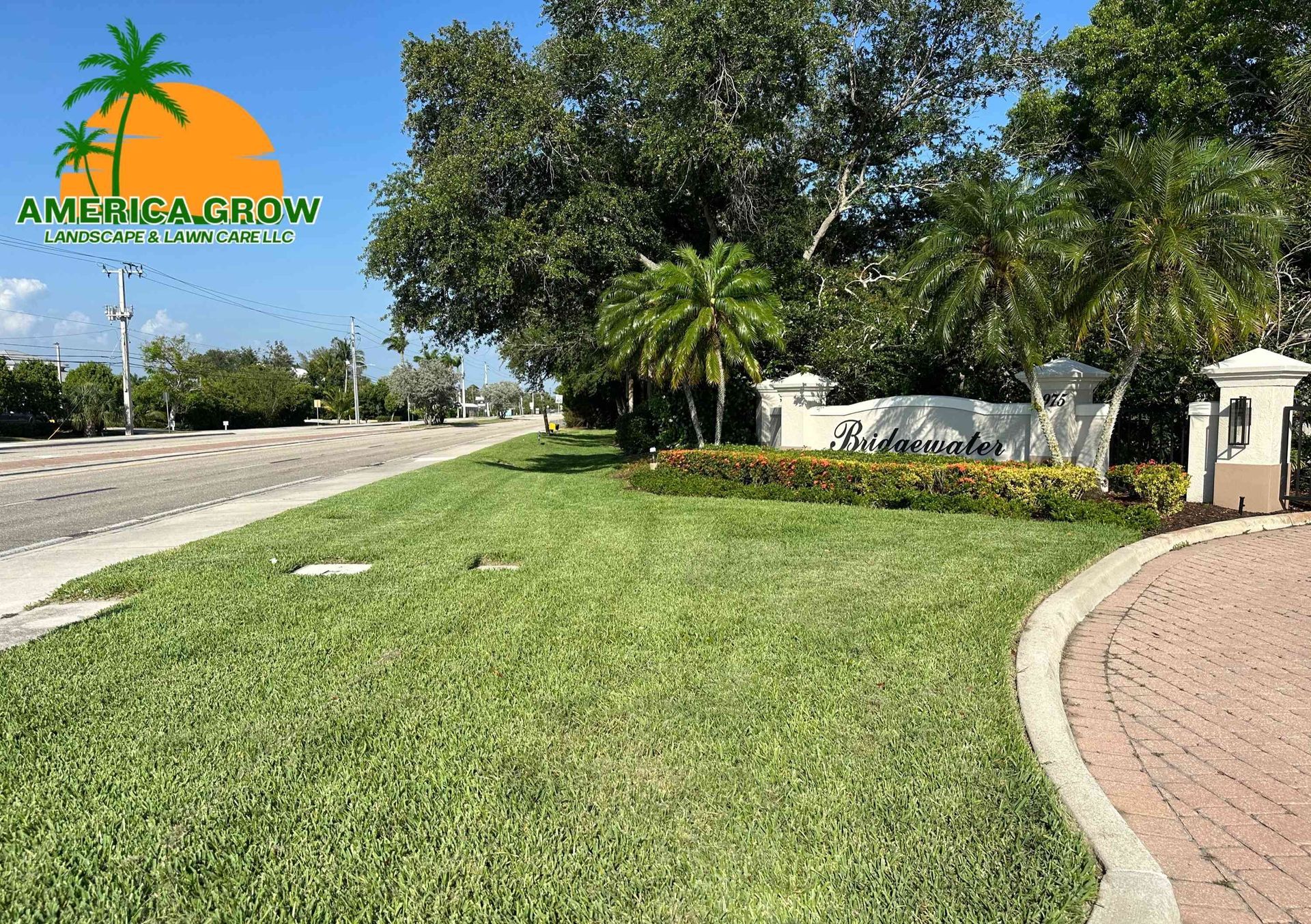 Lush green lawn with a palm tree logo, leading to a community entrance with a street and trees in the background.
