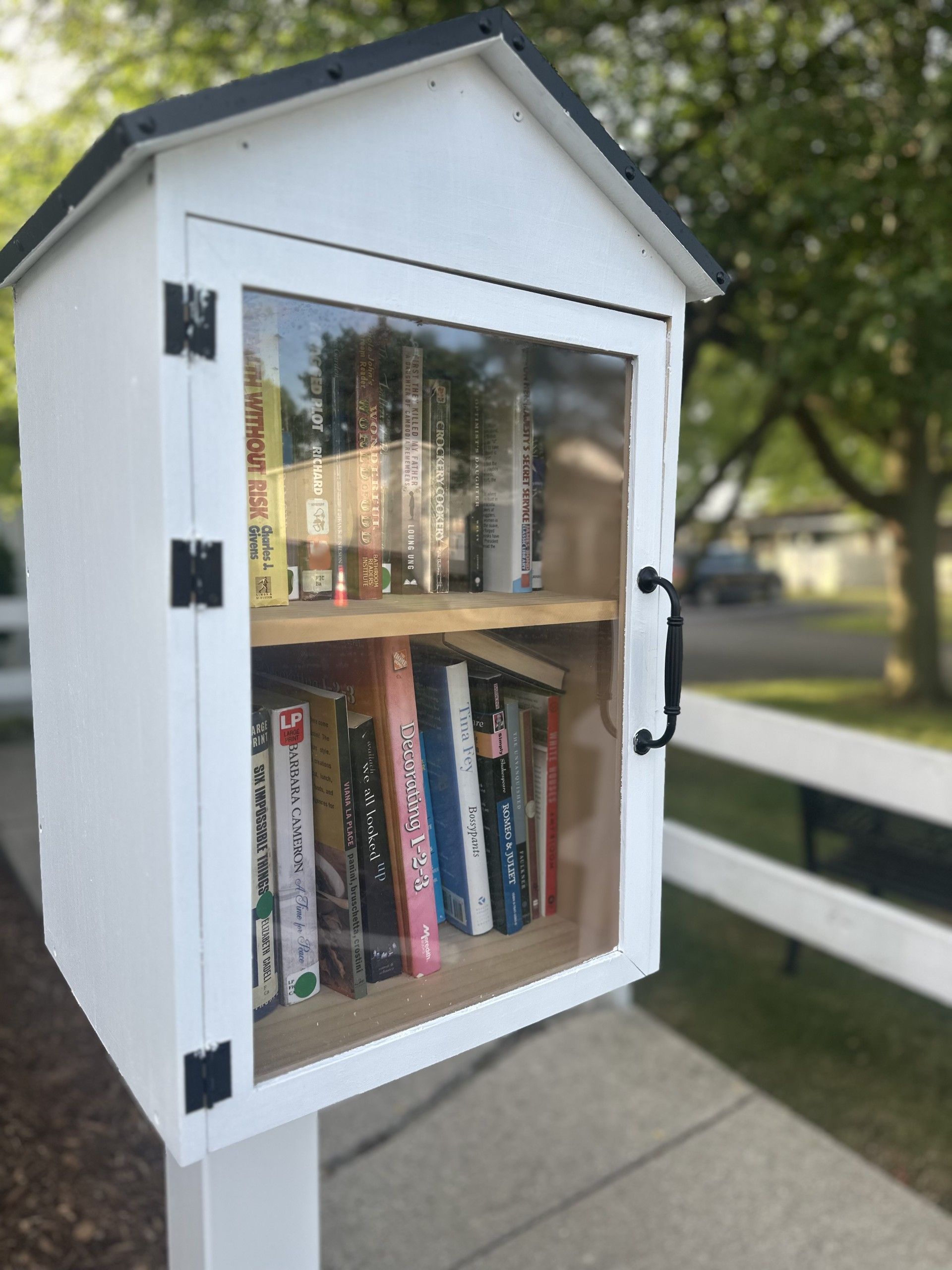 White Little Free Library on a post, filled with books. Black door hardware, sunny outdoor setting.