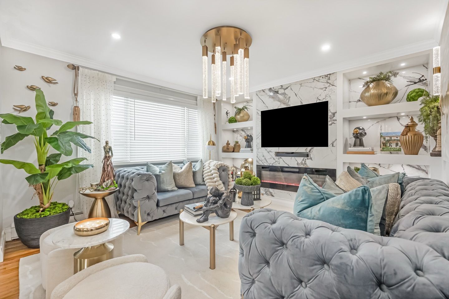 Bright modern living room with tufted gray sofas, teal pillows, wall TV, and large windows with sheer curtains