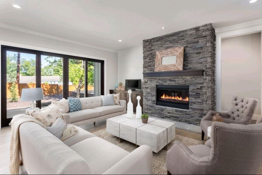Bright modern living room with white sofas, stone fireplace, and large windows overlooking greenery