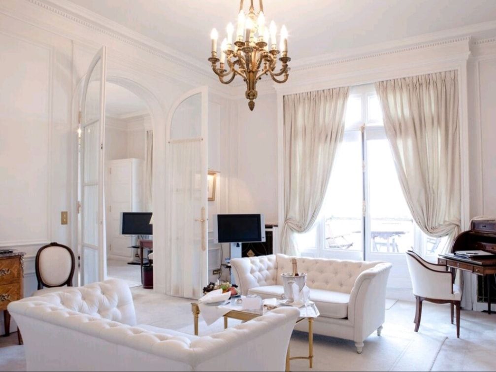 Elegant white living room with chandelier, arched mirrors, sofas, and tall curtains by a bright window