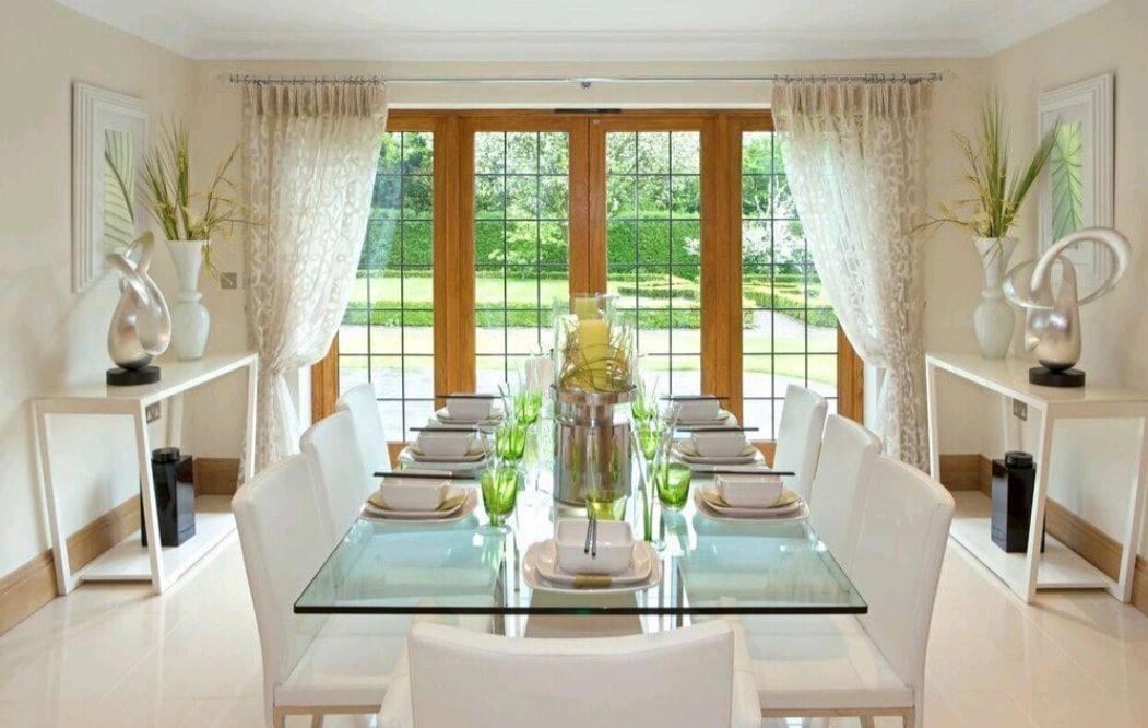Elegant dining room with a glass table, white chairs, and large windows overlooking a green garden
