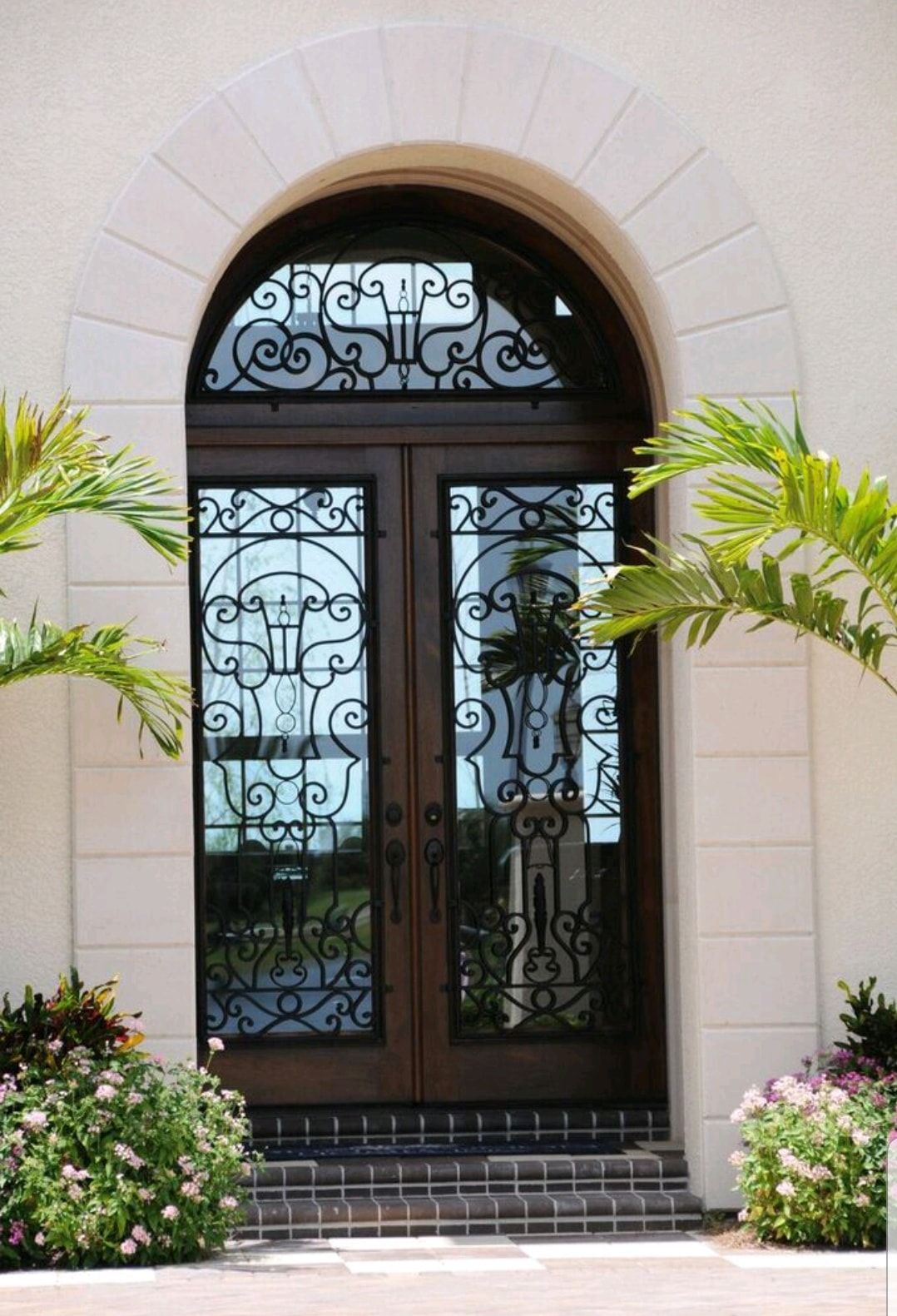 Arched front doorway with ornate black wrought-iron glass doors, framed by potted plants and white stone trim