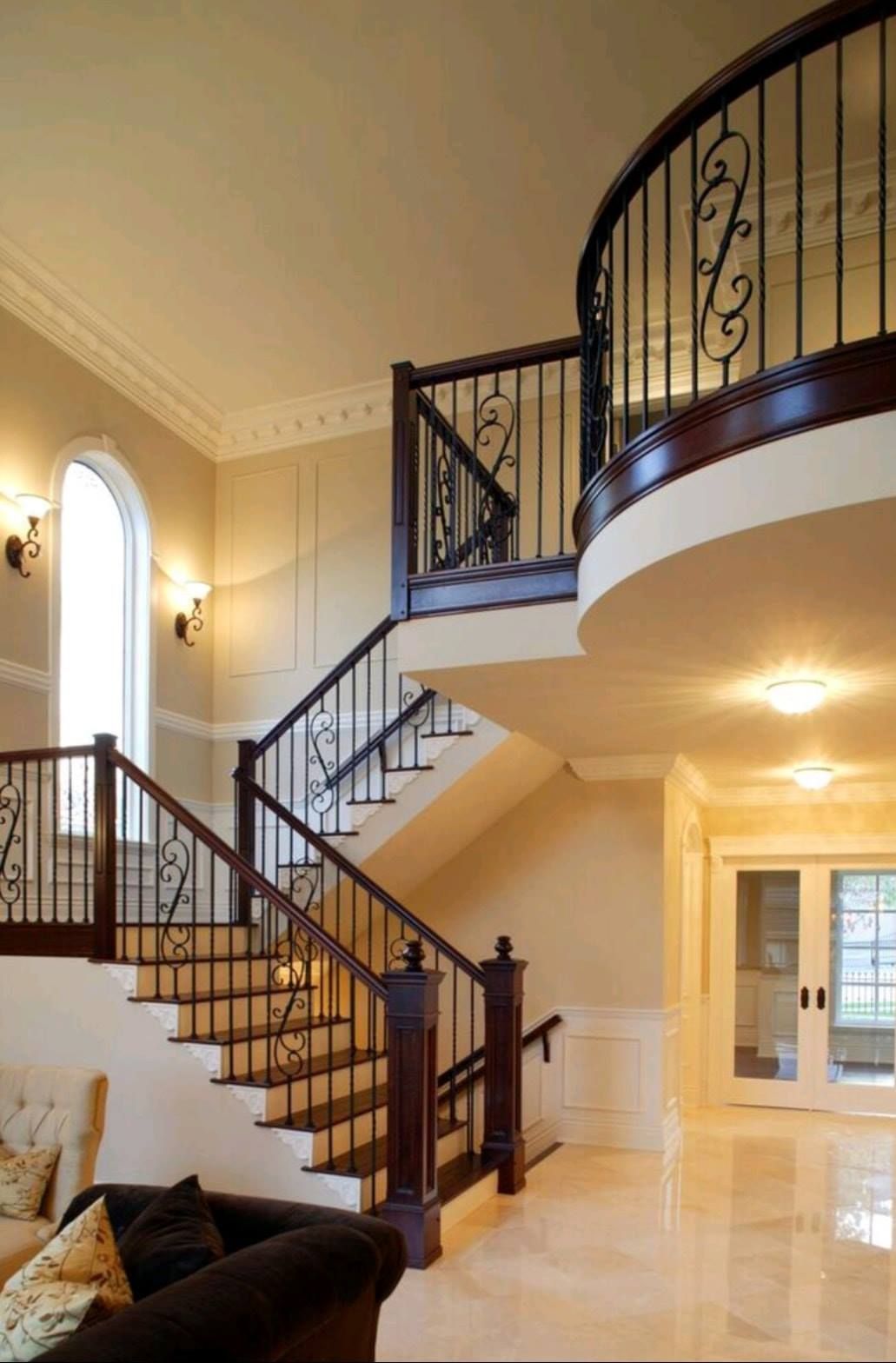 Elegant foyer with curved staircase, black railings, marble floor, and arched balcony under warm lighting