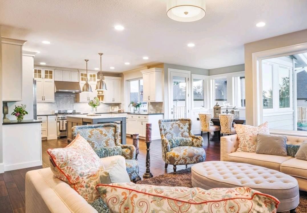 Bright living room with floral sofas, tufted ottoman, and open kitchen with white cabinets and large windows